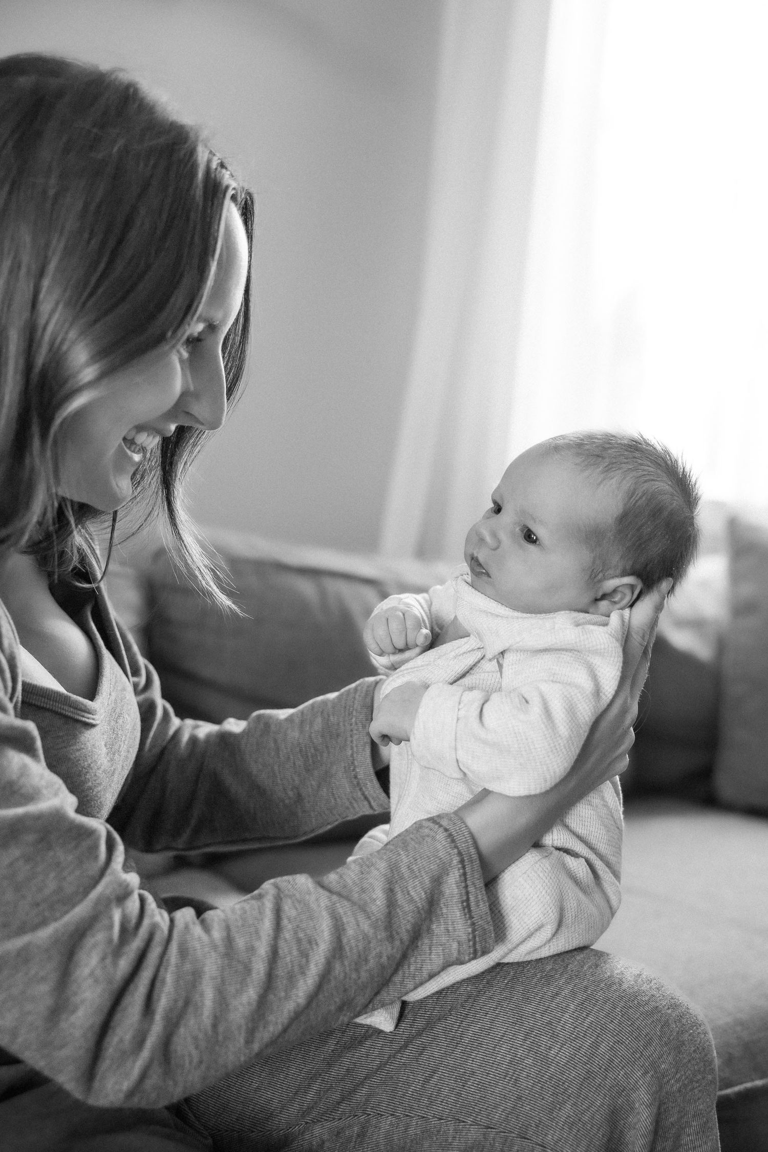 A smiling mother holds her alert newborn baby in a bright living room in a black and white photo.