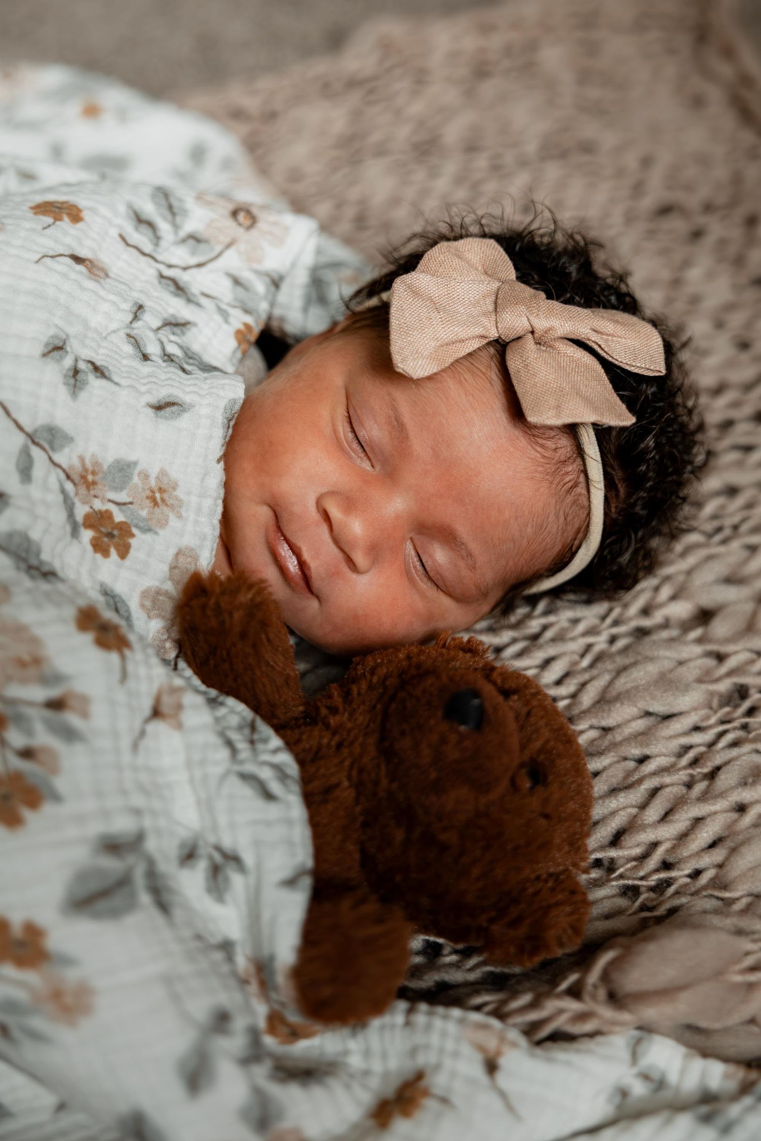 Sleeping newborn baby girl wearing a bow headband, wrapped in a floral swaddle, cuddling a brown teddy bear.