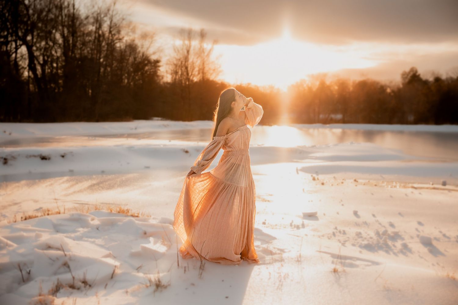 Woman in flowing peach gown standing in snowy landscape during golden sunset with trees in background.Lifestyle Virginia Photographer Anglin