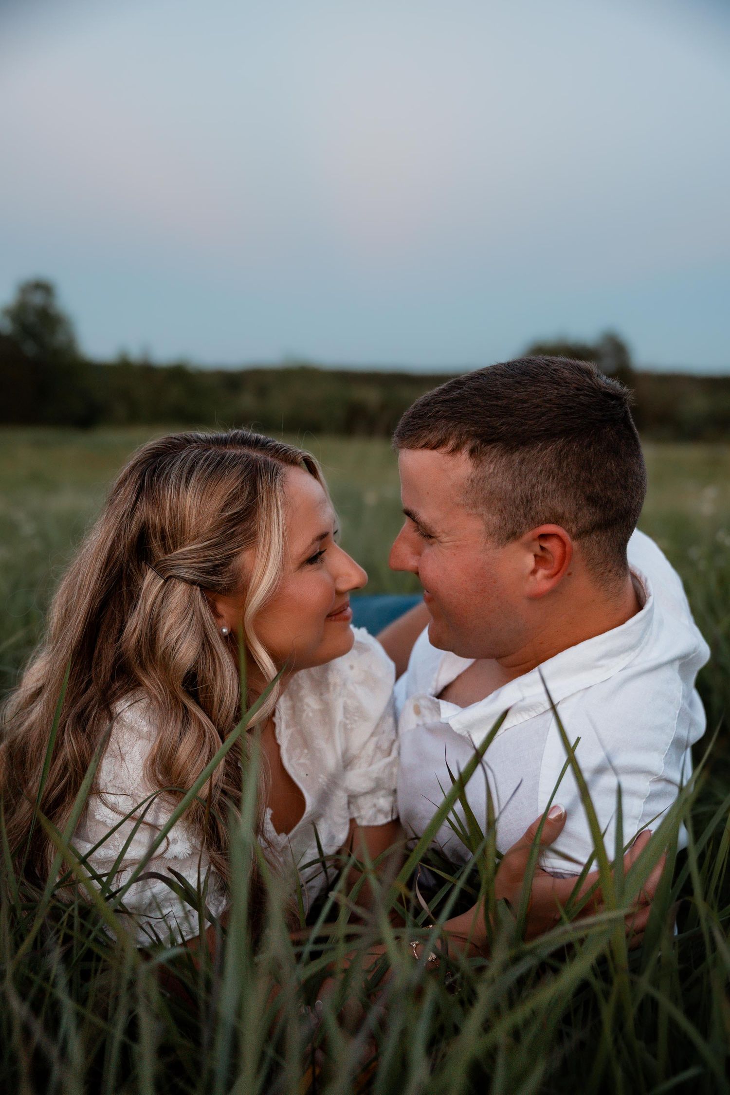A couple sitting in tall grass in a field, gazing lovingly at each other during golden hour.