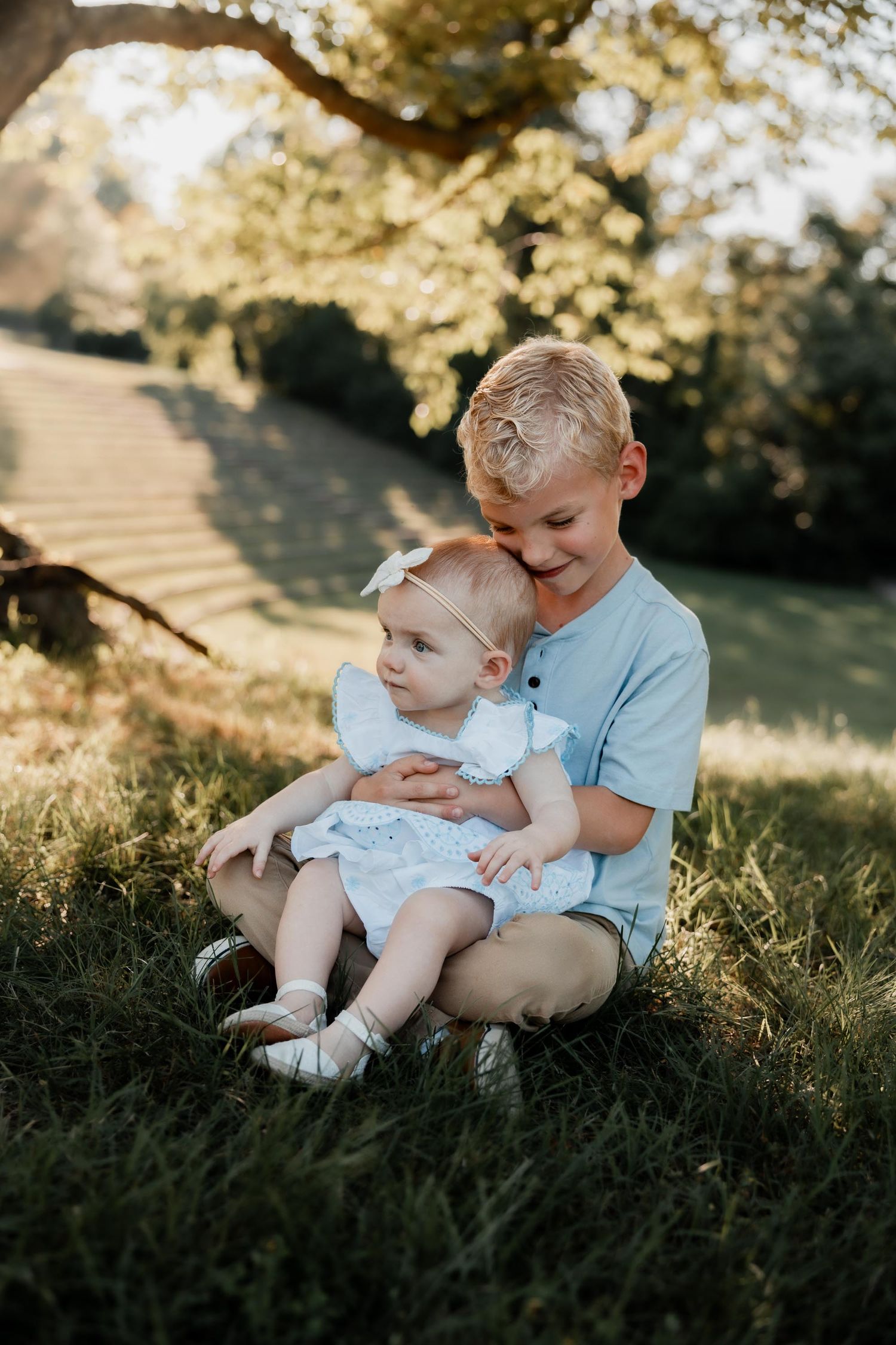 Older blonde boy sitting on grass cradling baby girl in white dress outdoors in golden sunlight.