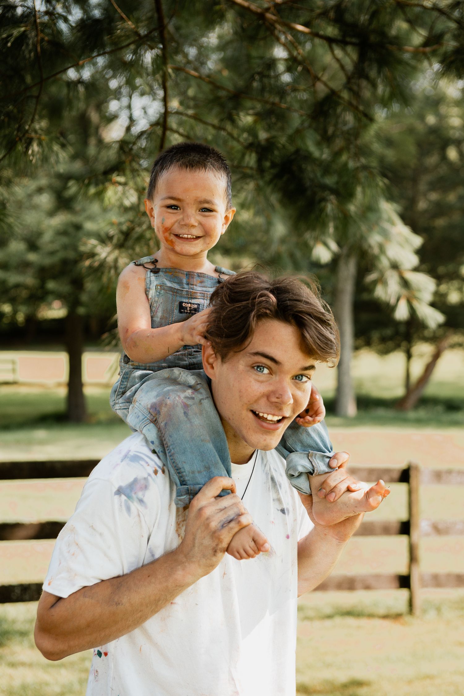 A playful outdoor moment shows a shoulder carry ride beneath green pine branches on a sunny day.