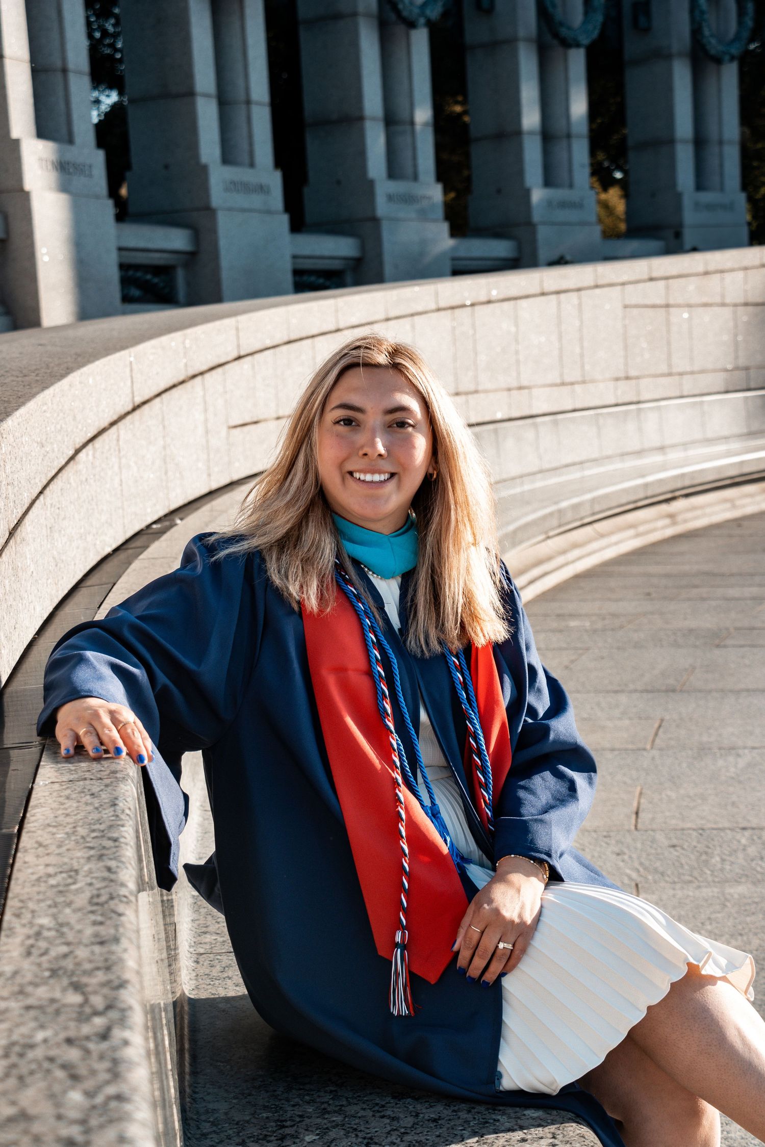 College graduate in navy graduation gown and red stole sits smiling on white stone steps of university building.