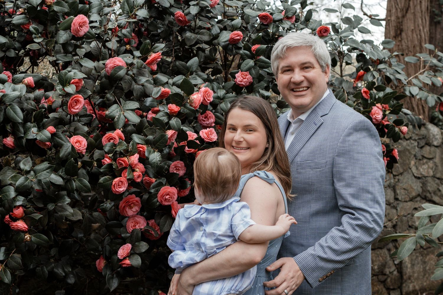 Family poses together against vibrant red camellia flowers blooming on dark green bushes in garden setting.
