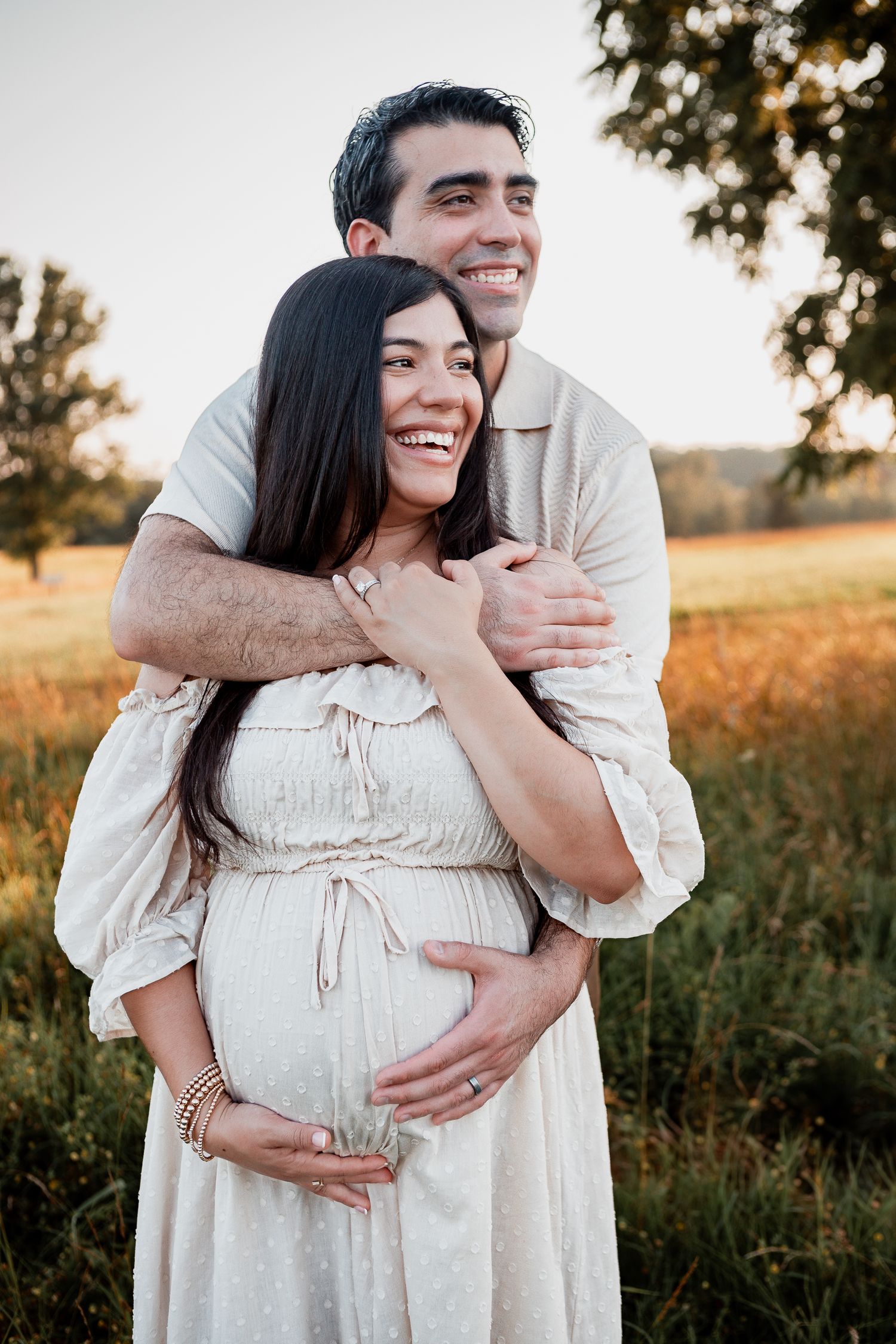 A couple embraces in a golden field at sunset while expecting their first child together.