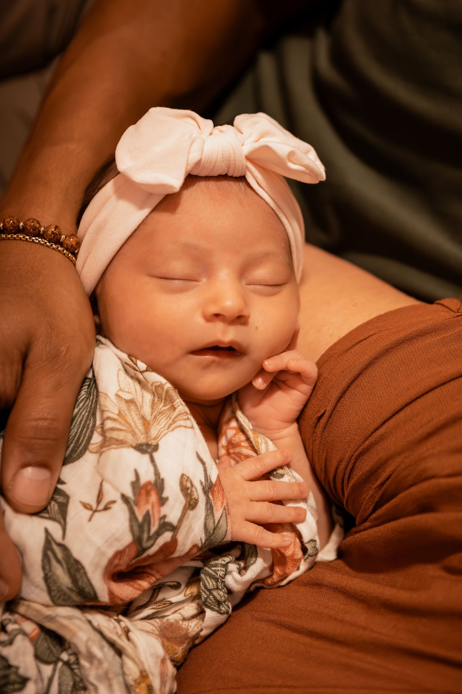 Adorable sleeping infant in floral onesie rests peacefully on brown fabric with delicate lighting creating a warm atmosphere.