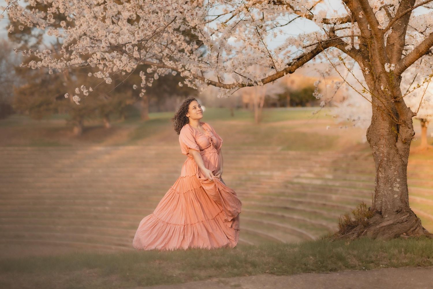 Woman in flowing peach gown standing under blooming cherry blossom tree in golden hour light.