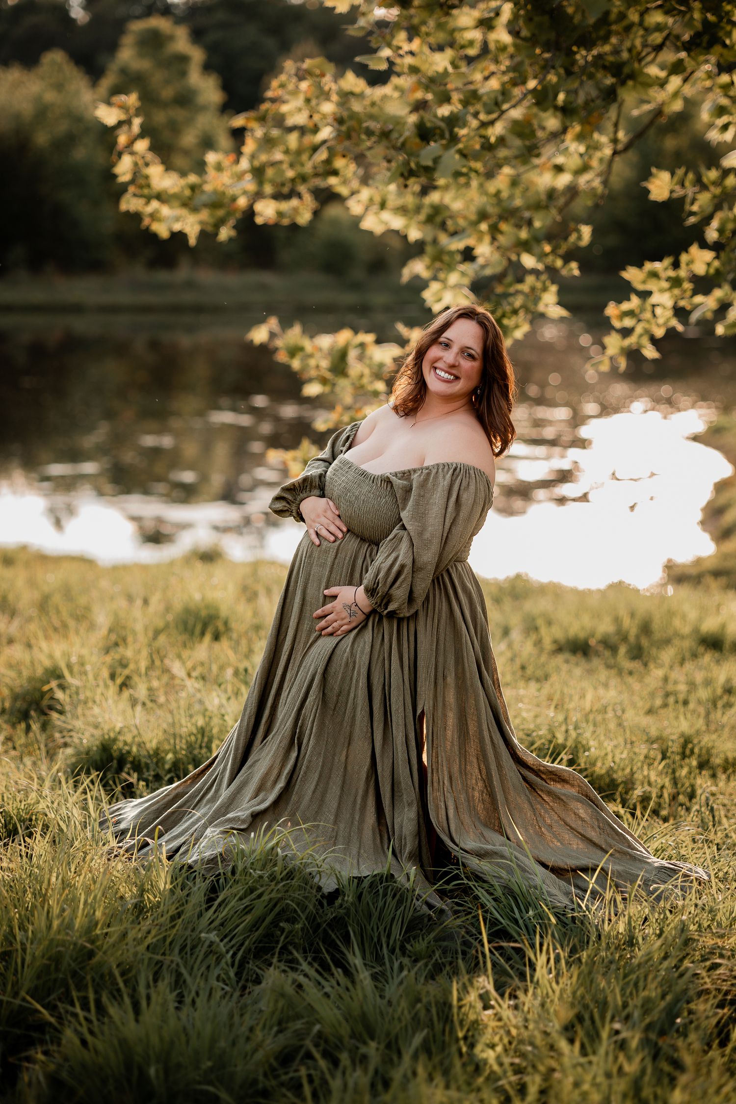 Smiling pregnant woman in olive green off-shoulder gown poses outdoors near a pond at golden hour.