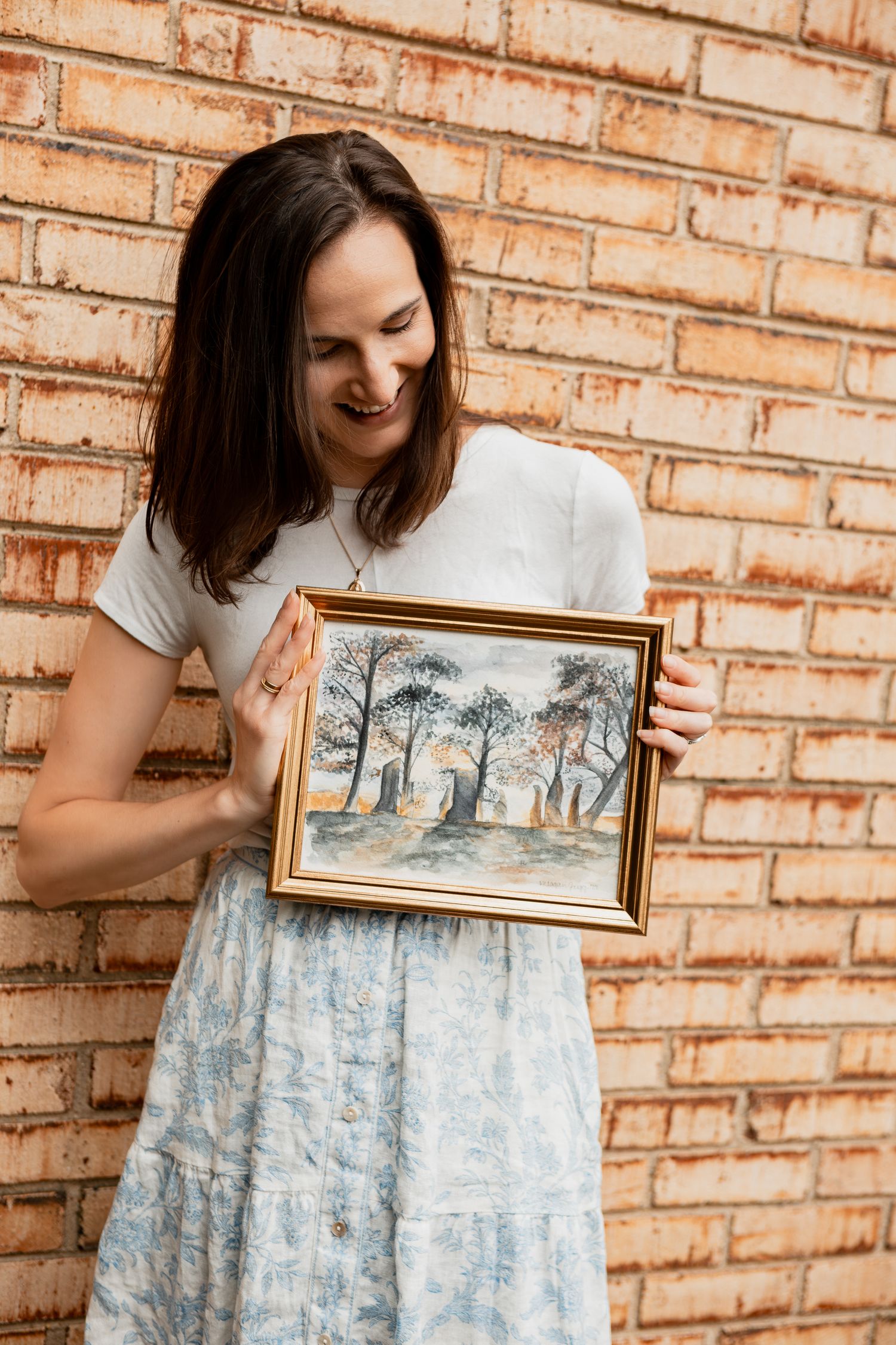 Woman in white top holds framed landscape artwork while standing against brick wall.