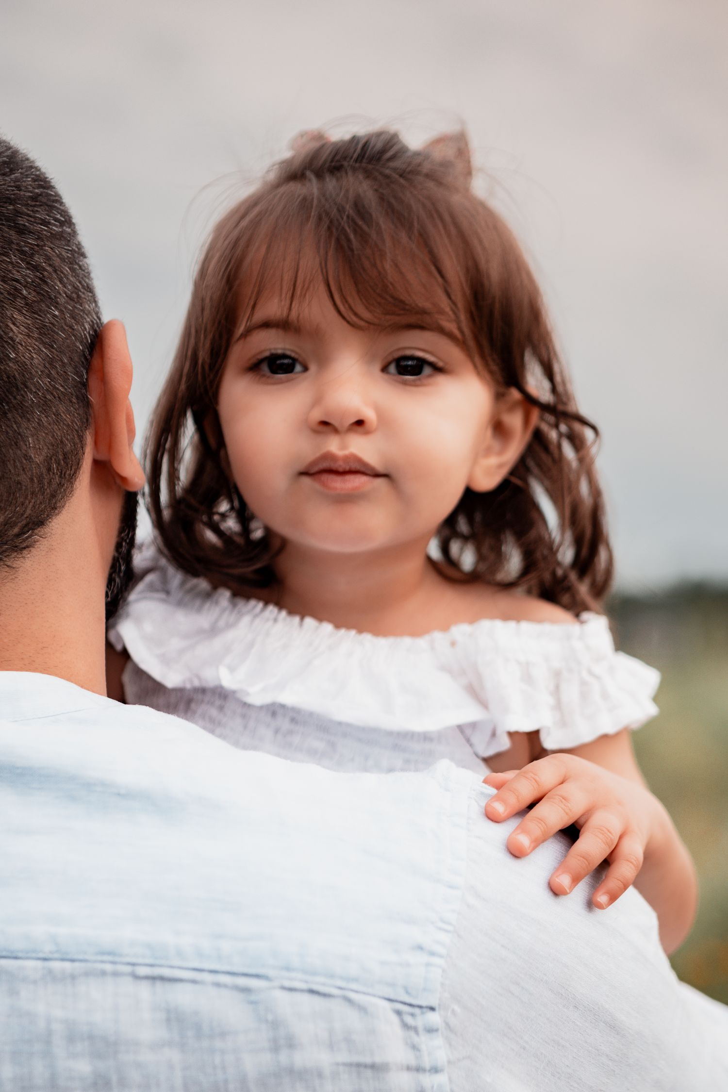 Child with pigtails in a white ruffle dress looks over someone's shoulder in an outdoor portrait.