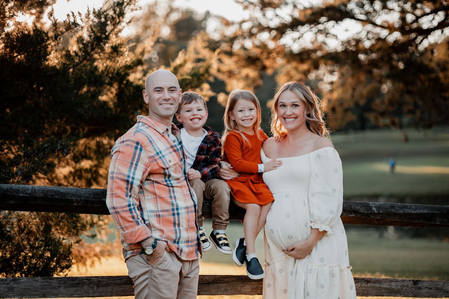 A family poses together outdoors at sunset with golden light filtering through trees in the background.