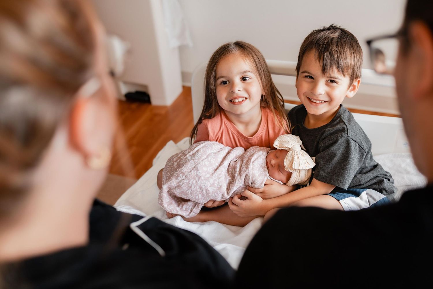 Two smiling children proudly hold a swaddled newborn baby while sitting on a hospital bed with parents nearby.