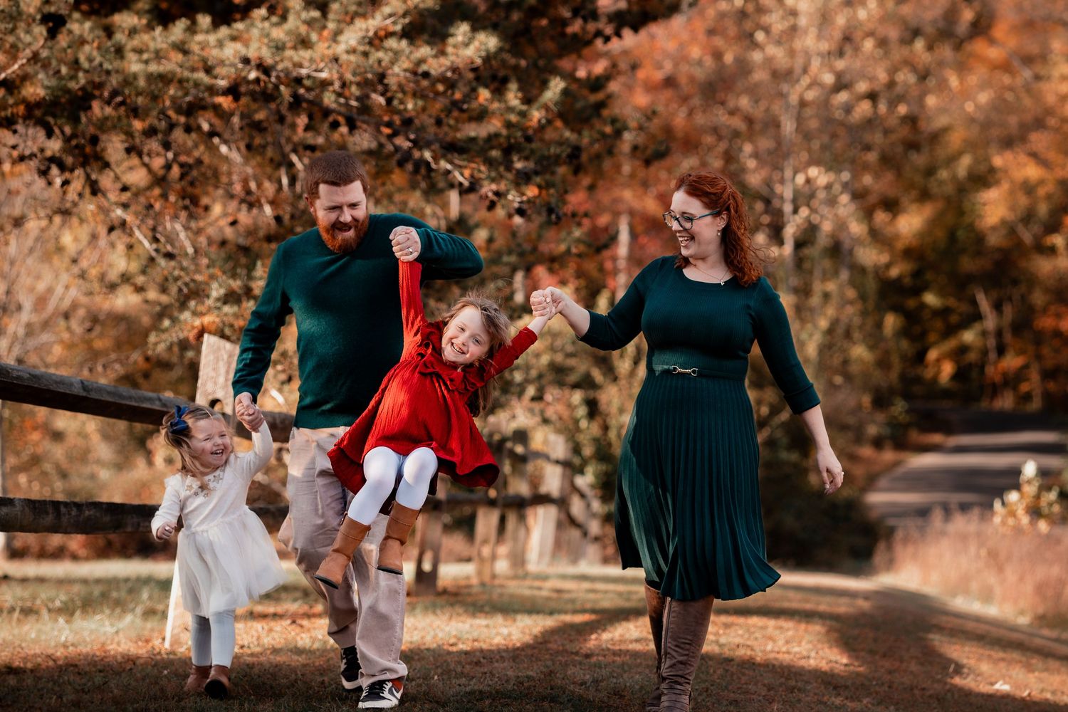 Family of four enjoying autumn walk, parents swinging happy children outdoors near wooden fence with fall foliage backdrop.