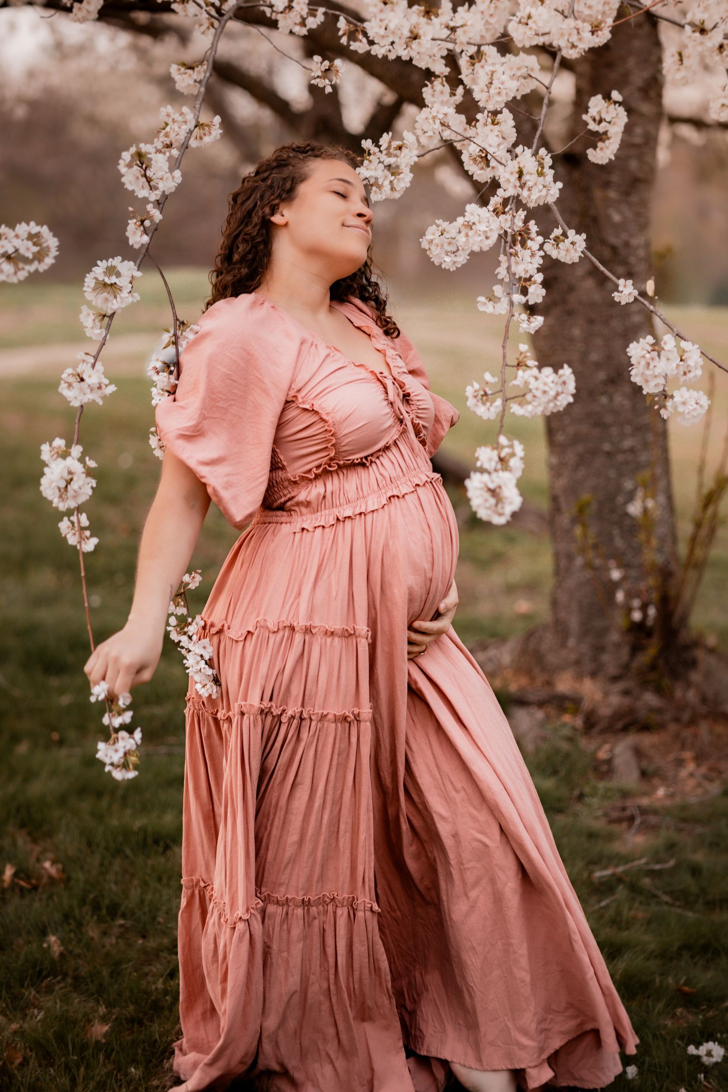 Pregnant woman in pink maxi dress smiling among cherry blossoms in spring garden.