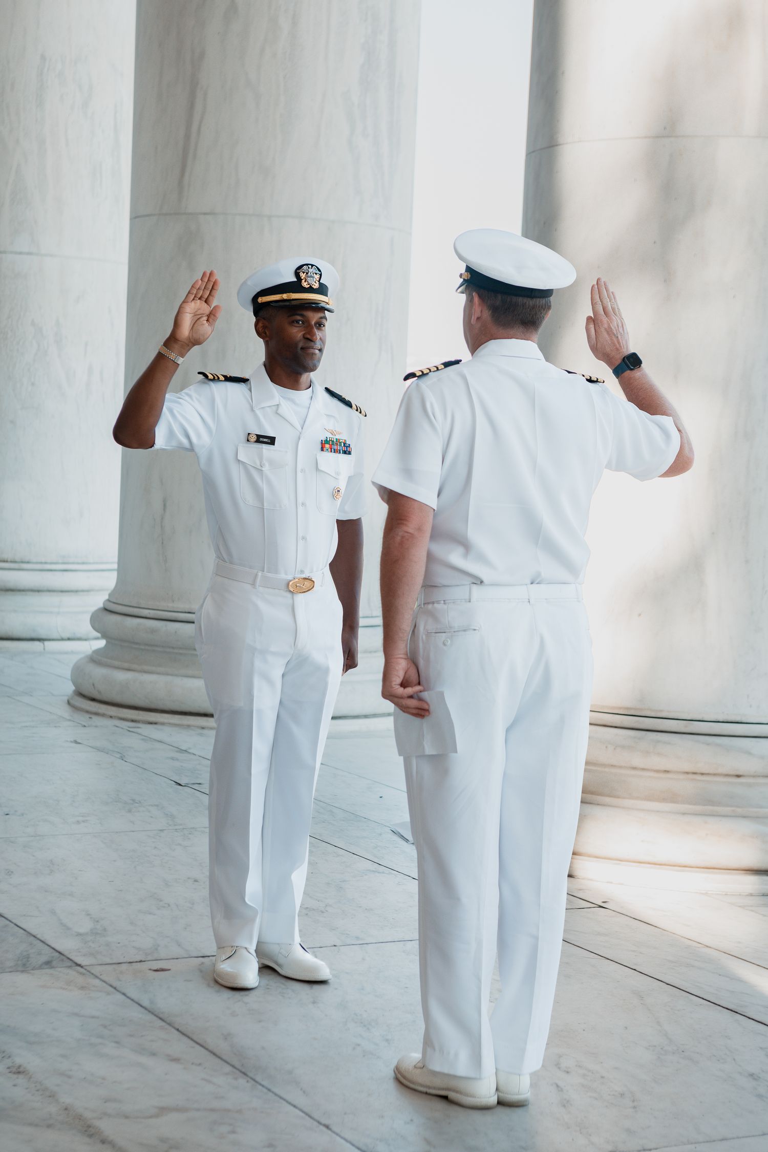 Two naval officers in white uniforms stand in salute position near classical columns in bright sunlight.