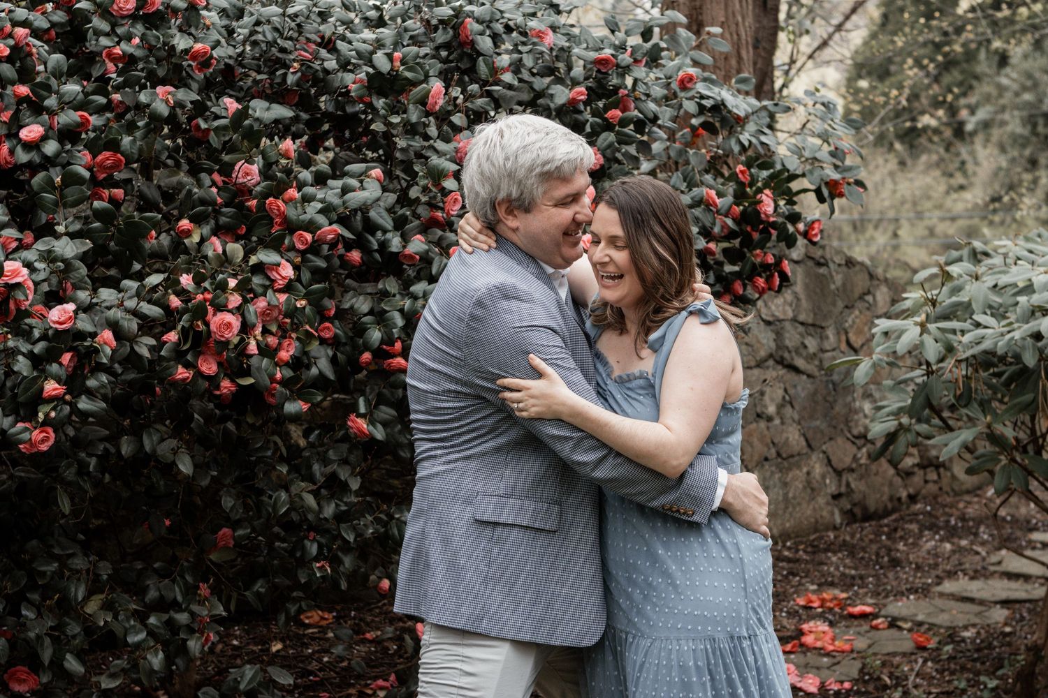 A romantic couple embraces in front of a blooming red camellia bush on a moody afternoon.