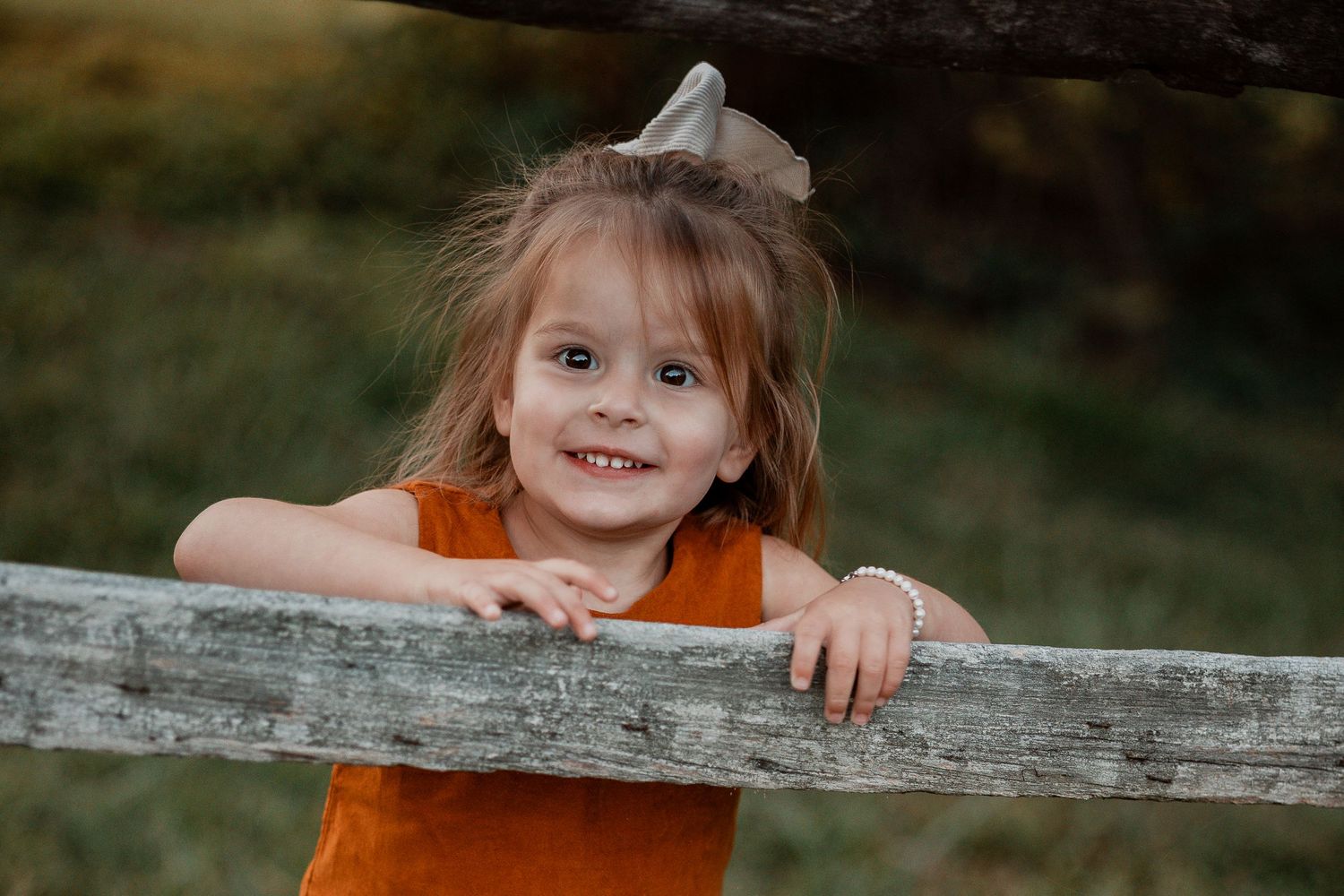 A child in an orange shirt peeks playfully over a rustic wooden fence in an outdoor setting.