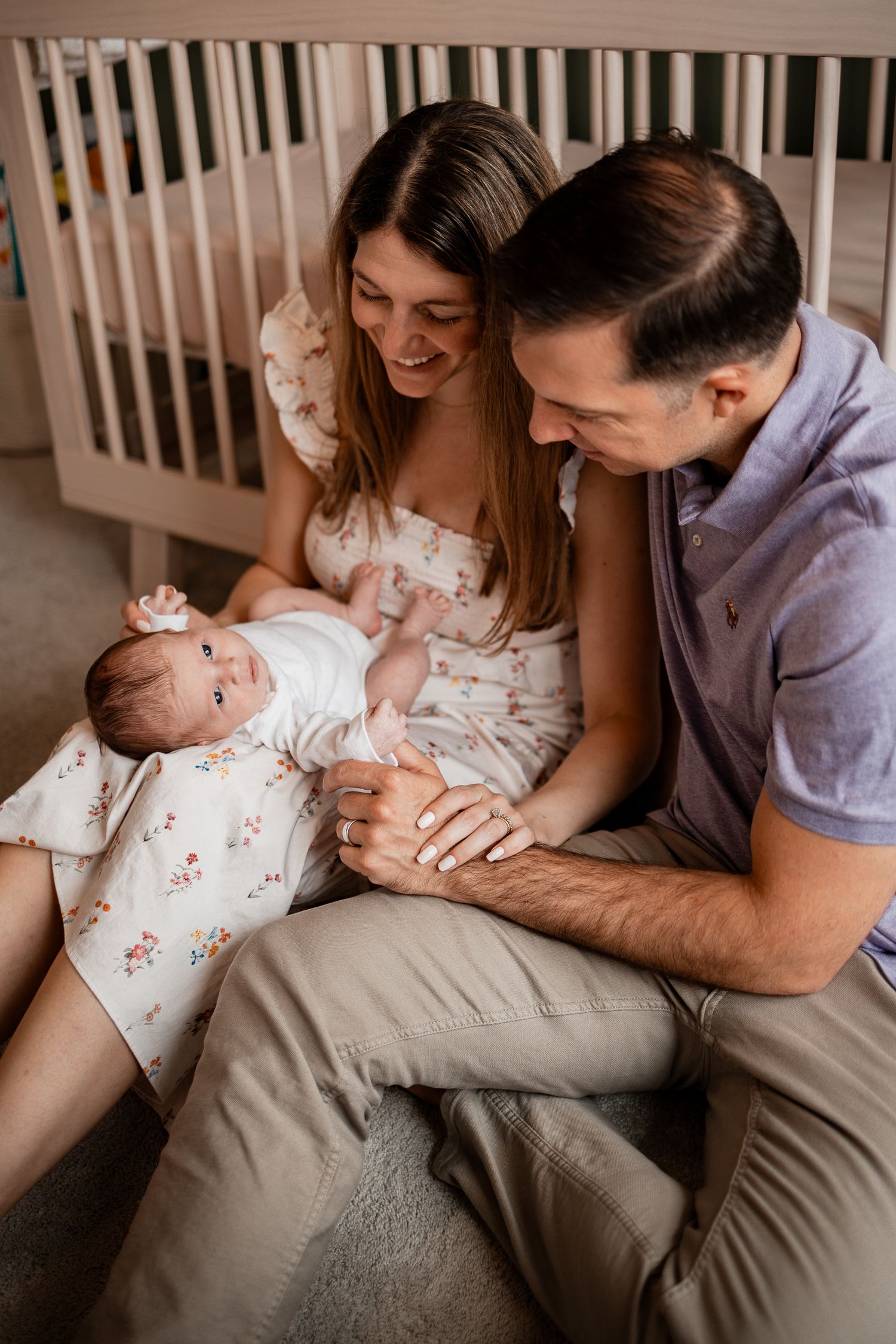 A couple sits lovingly with their newborn baby in a cozy nursery setting beside a white crib.