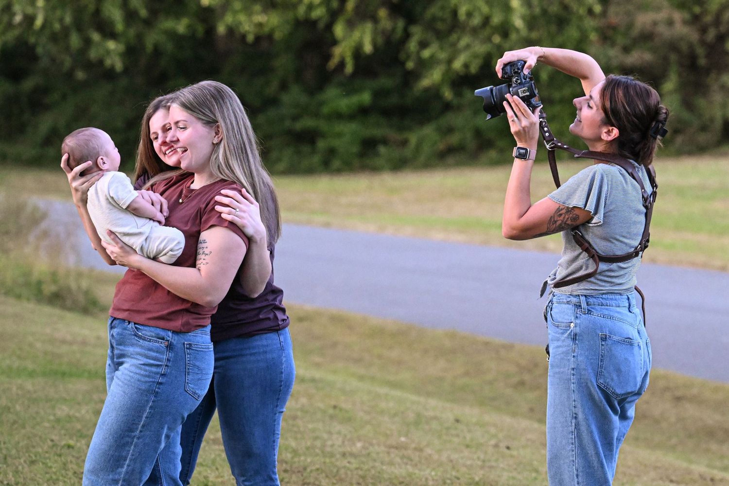 Photographer capturing two women holding a baby outdoors on a grassy field near a road.