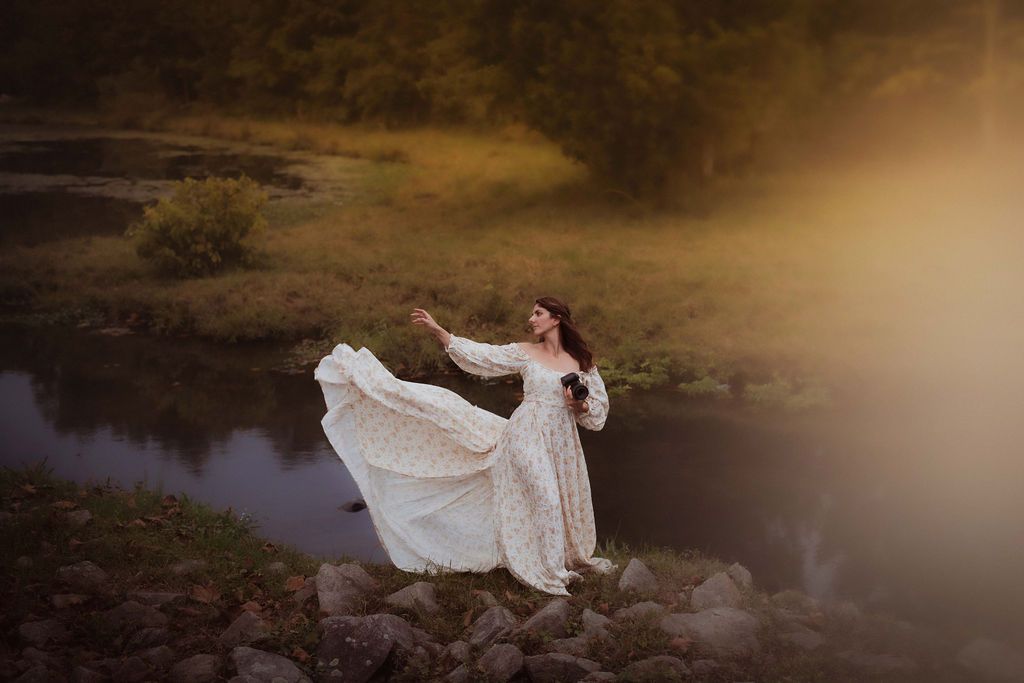Woman in white lace gown twirling on rocks by a misty river at golden hour in a dreamy outdoor setting.
