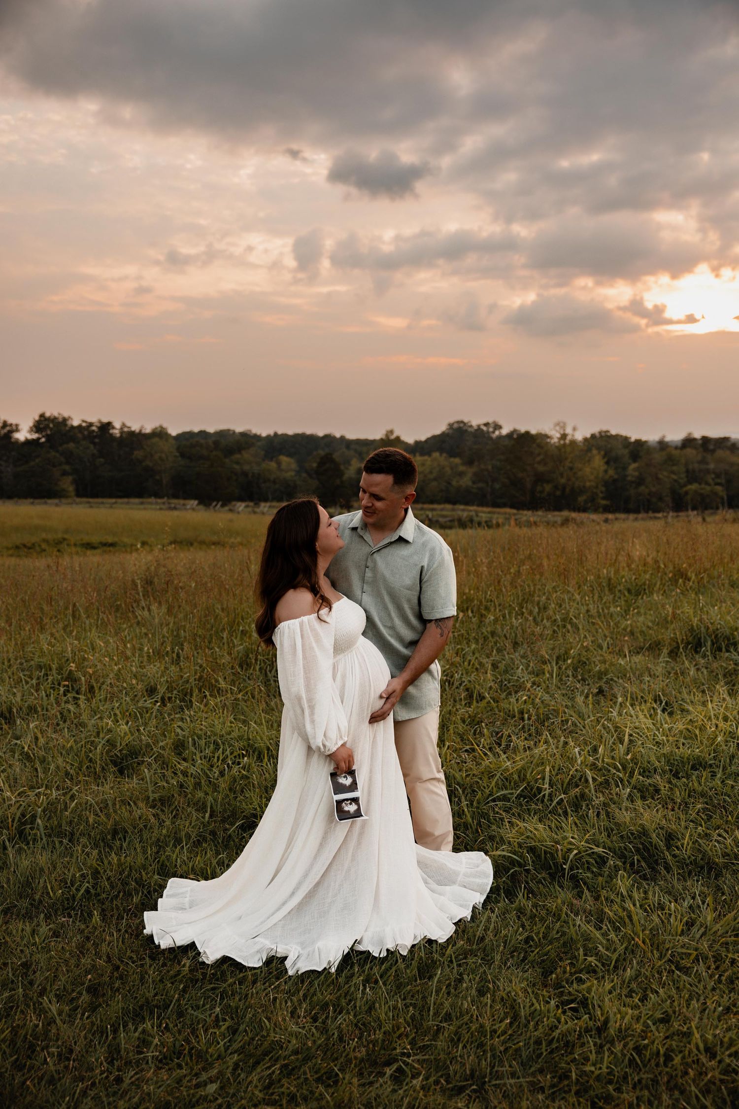 Expecting couple holding ultrasound photos while gazing at each other in open field at dramatic sunset.
