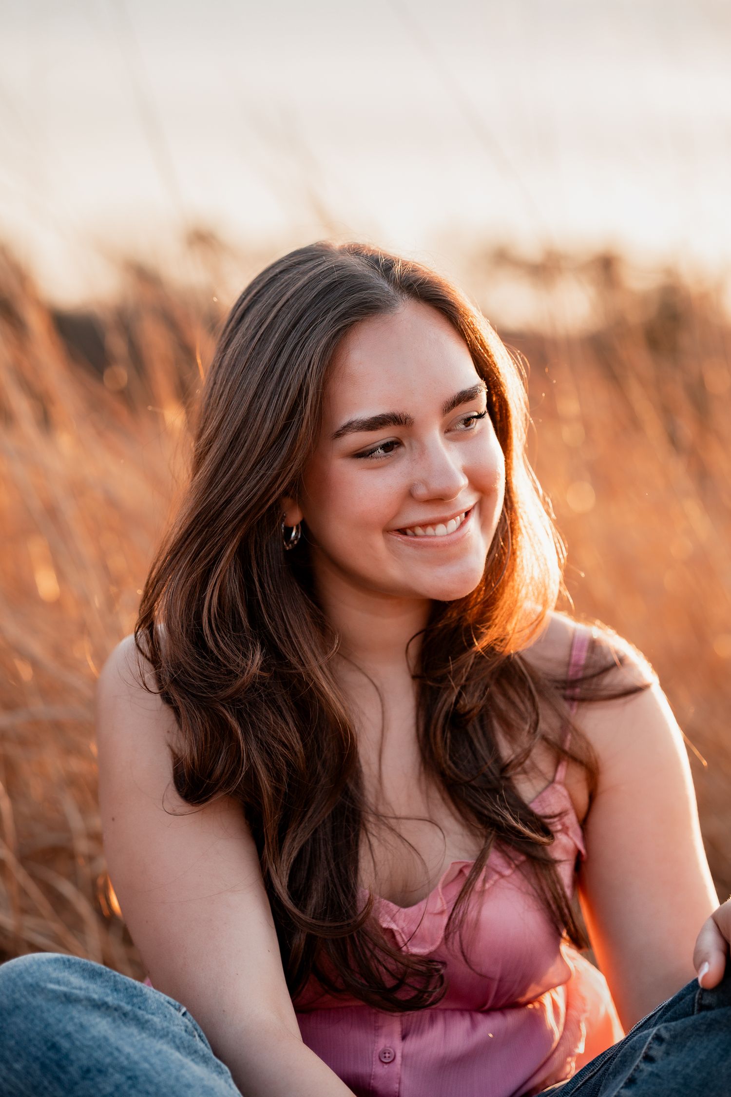 Young woman with long brown hair smiling outdoors in golden field at sunset, wearing pink top and jeans.