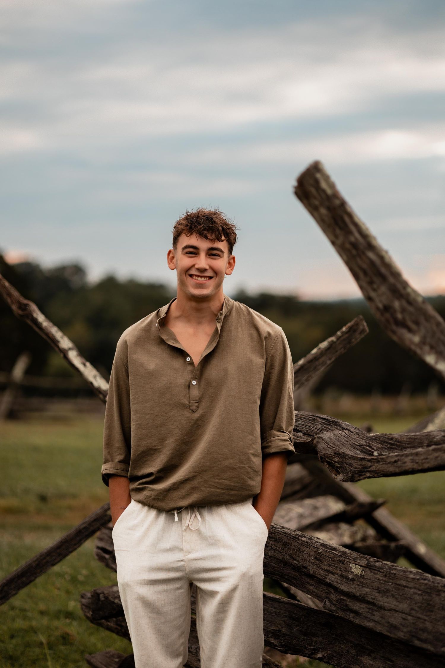 Young man in olive shirt and white pants smiling outdoors near rustic wooden fence in green field.