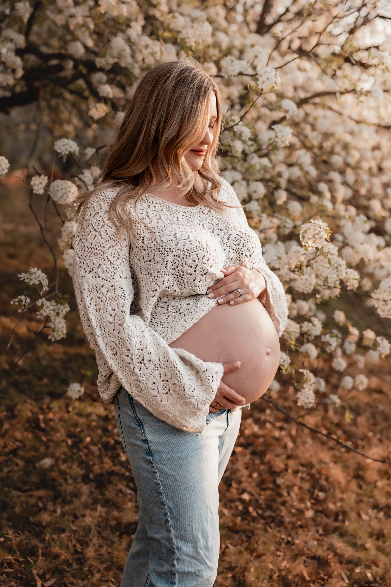 Pregnant woman in white lace top and jeans cradling her baby bump outdoors near blooming white spring flowers.
