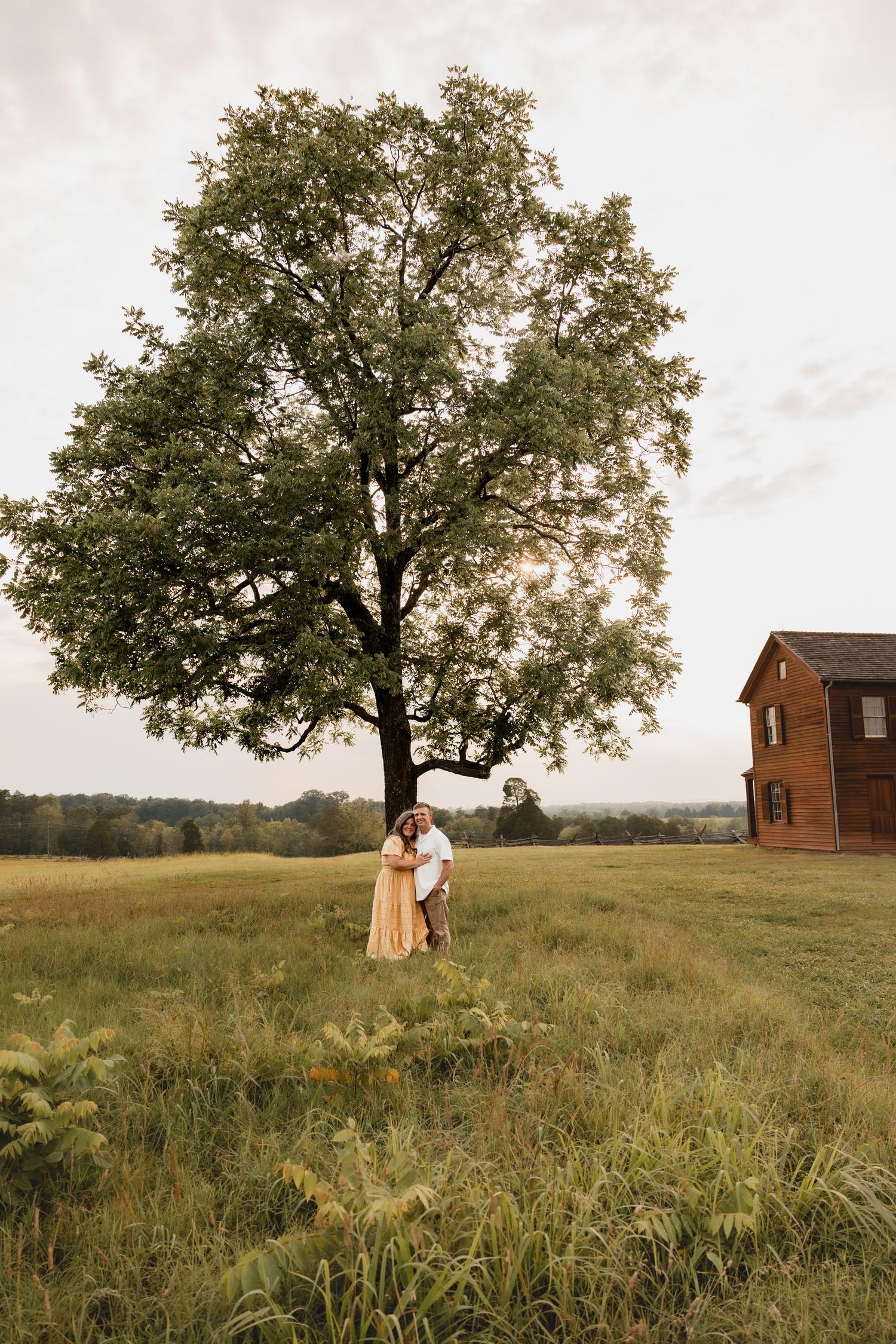 A couple embraces near a large tree in a scenic green meadow with a historic brick building at sunset.