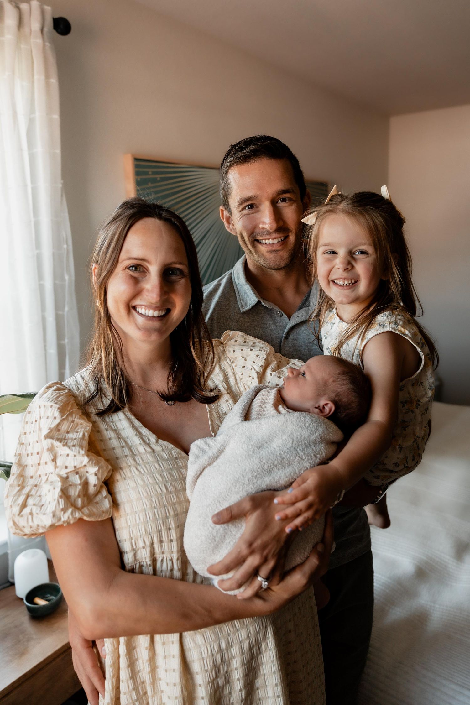 Happy family of four posing indoors, parents smiling while holding newborn baby and young daughter in bow.