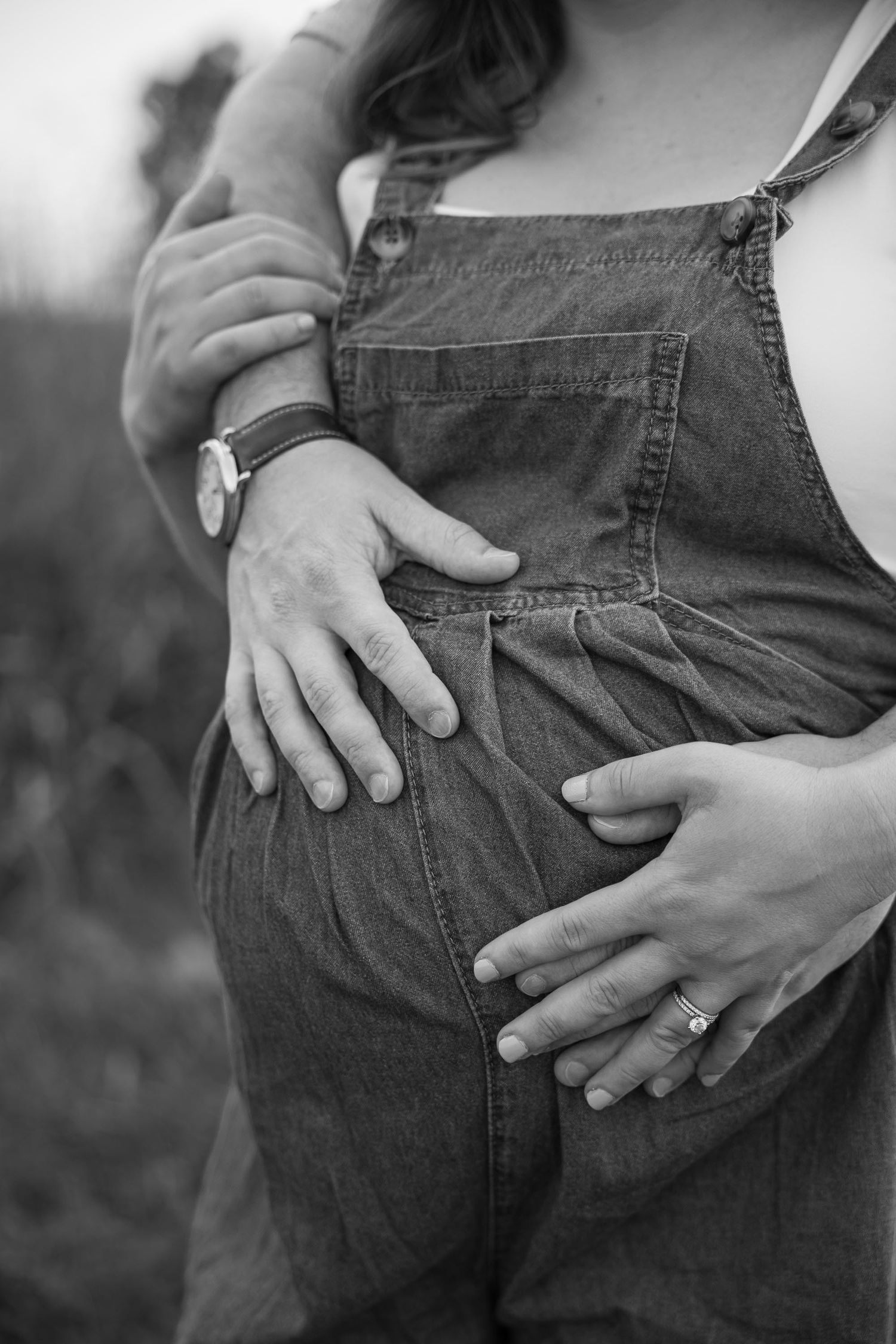 Black and white close-up of pregnant belly in denim overalls with couple's hands gently cradling the bump.