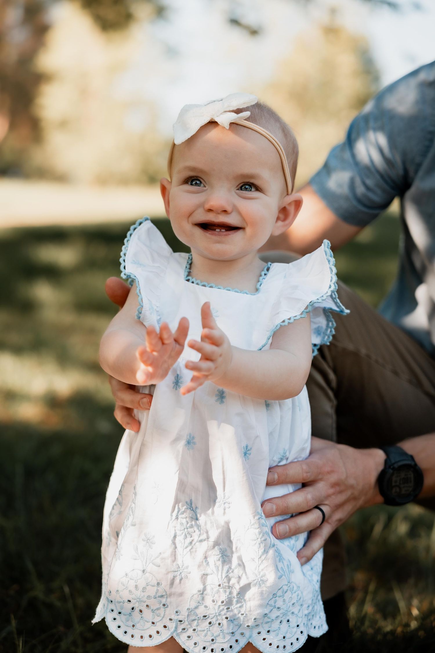 Smiling baby girl in white dress and bow headband clapping hands outdoors, held by adult in sunny park.