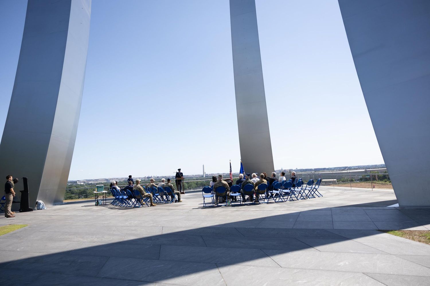 Outdoor ceremony at Air Force Memorial with tall silver spires under clear blue sky, attendees seated in blue chairs on stone plaza.