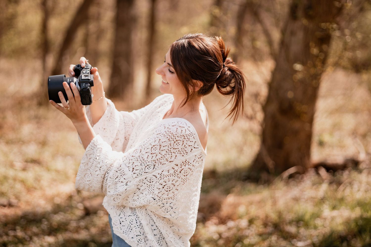 Young woman in white lace sweater holding a vintage camera outdoors in a wooded setting.