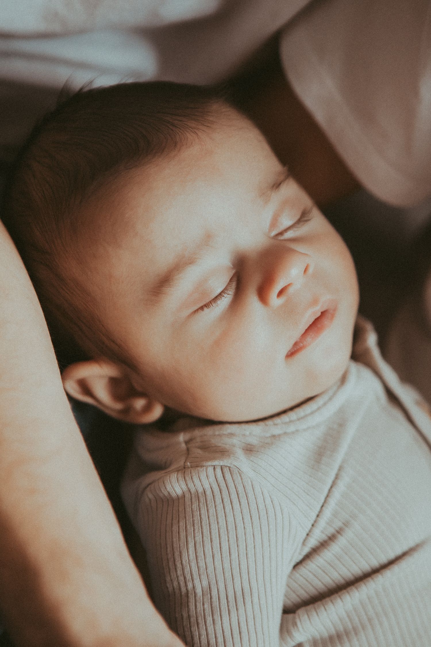 Sleeping infant wearing a soft cream-colored sweater in gentle natural lighting.