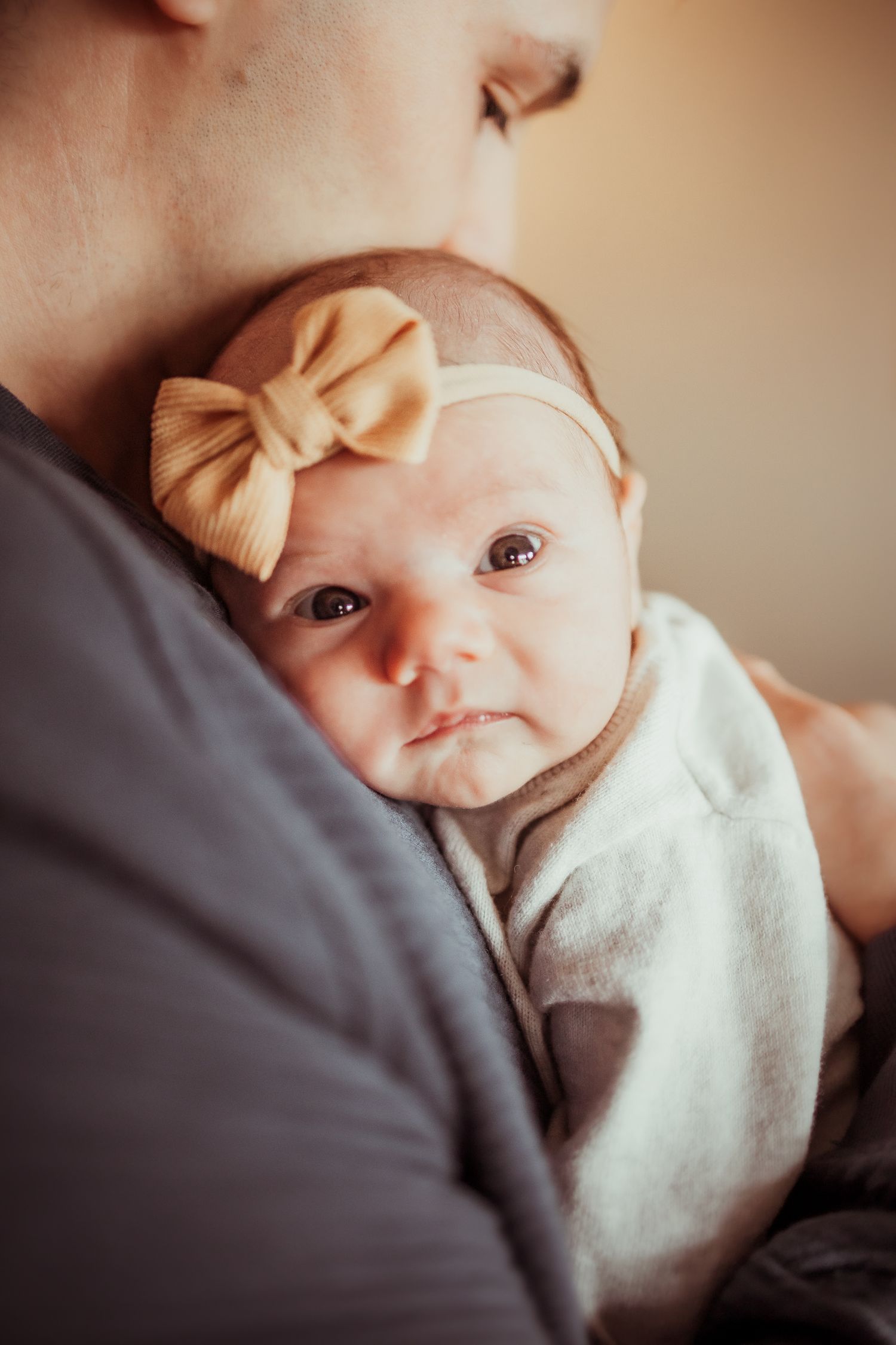 A baby wearing a cream bow headband and white swaddle blanket looks up at the camera with wide eyes.