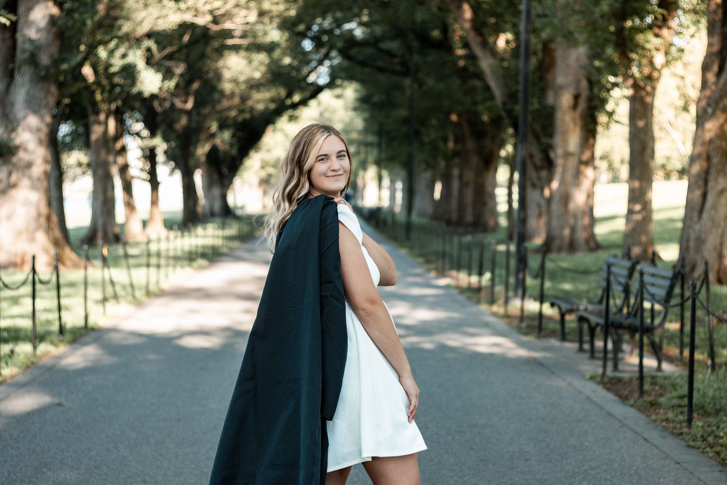 Bride in white sleeveless dress poses in a tree-lined path with sunlight filtering through branches.