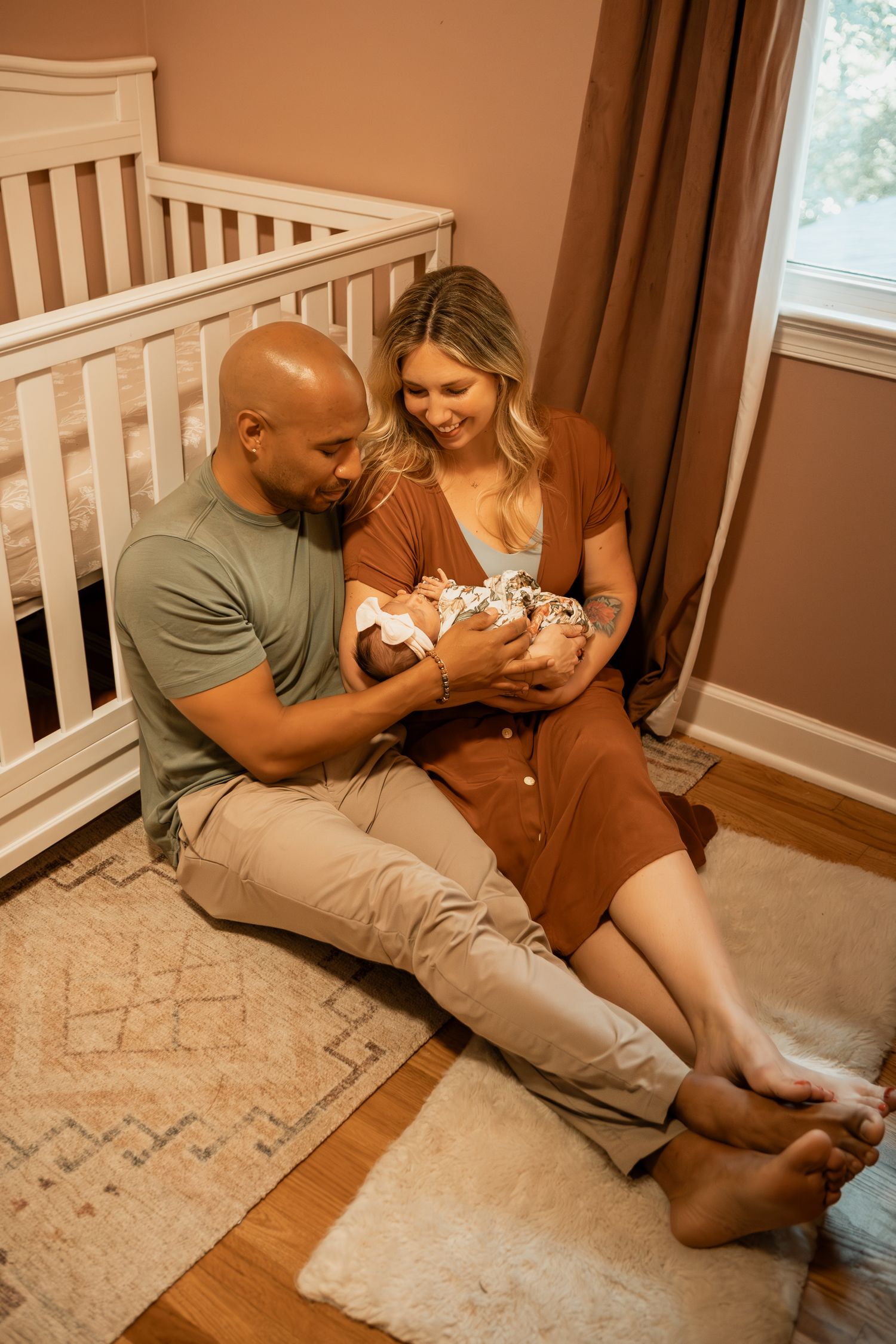 Parents in casual attire sitting together on floor with their newborn baby in nursery room with white crib visible.