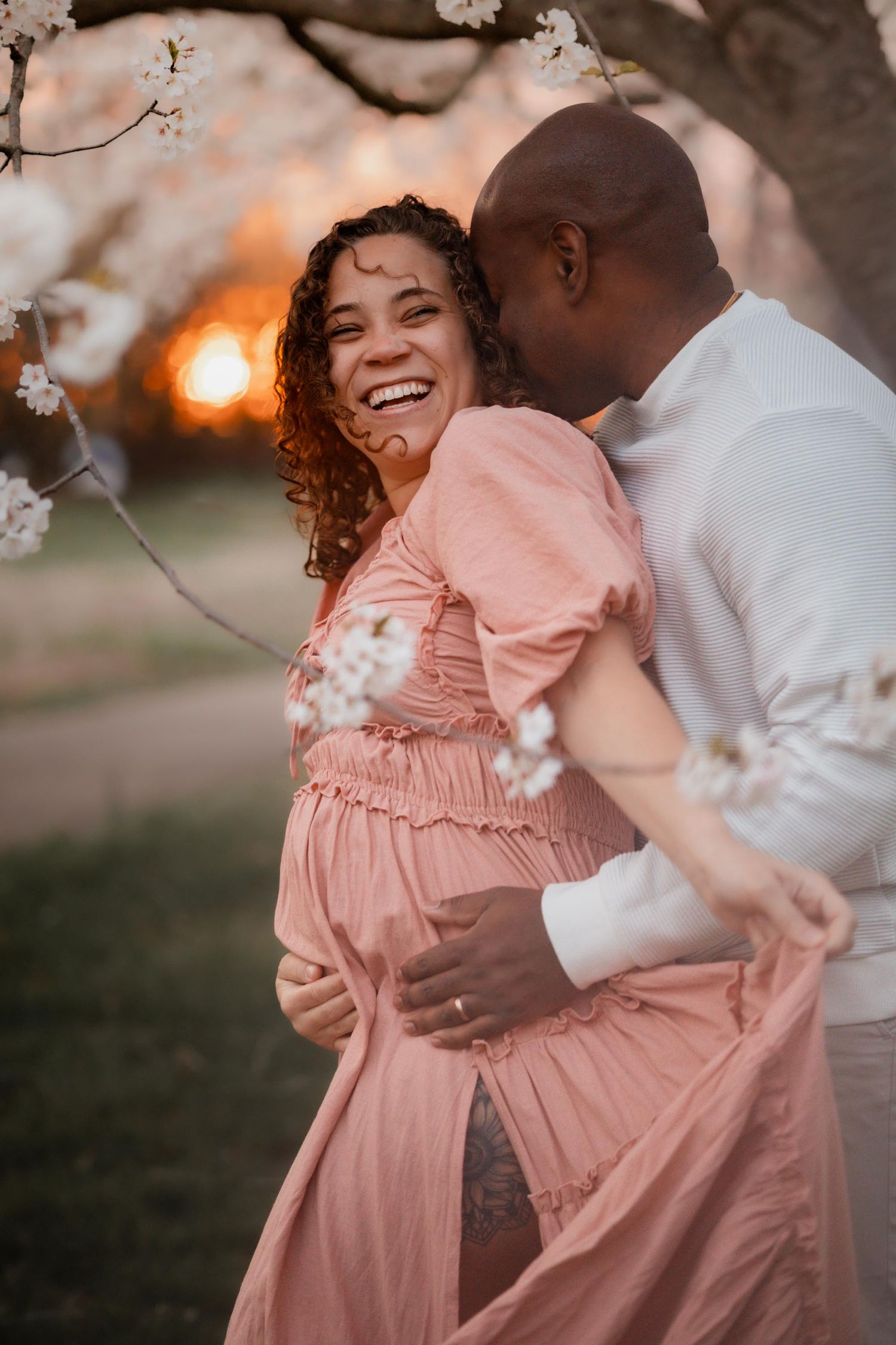 Pregnant woman in pink dress laughs joyfully as partner embraces her under cherry blossoms at golden hour.