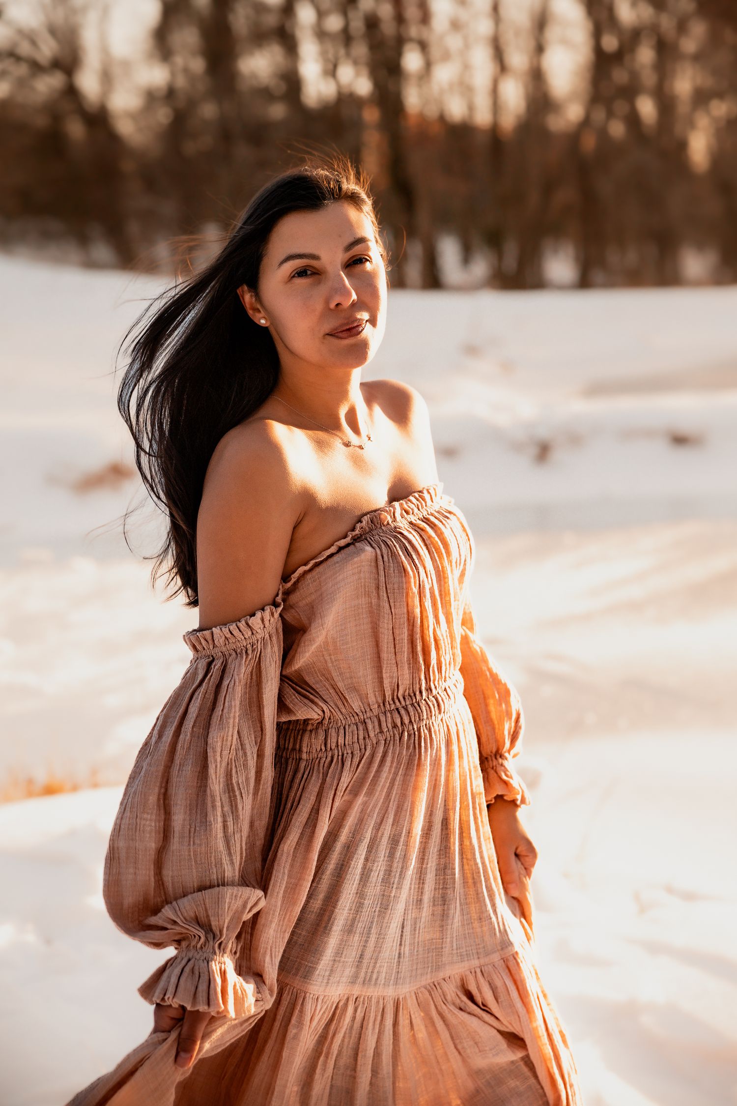 A dark-haired woman in an off-shoulder boho rust dress poses gracefully in a snowy winter landscape at golden hour.