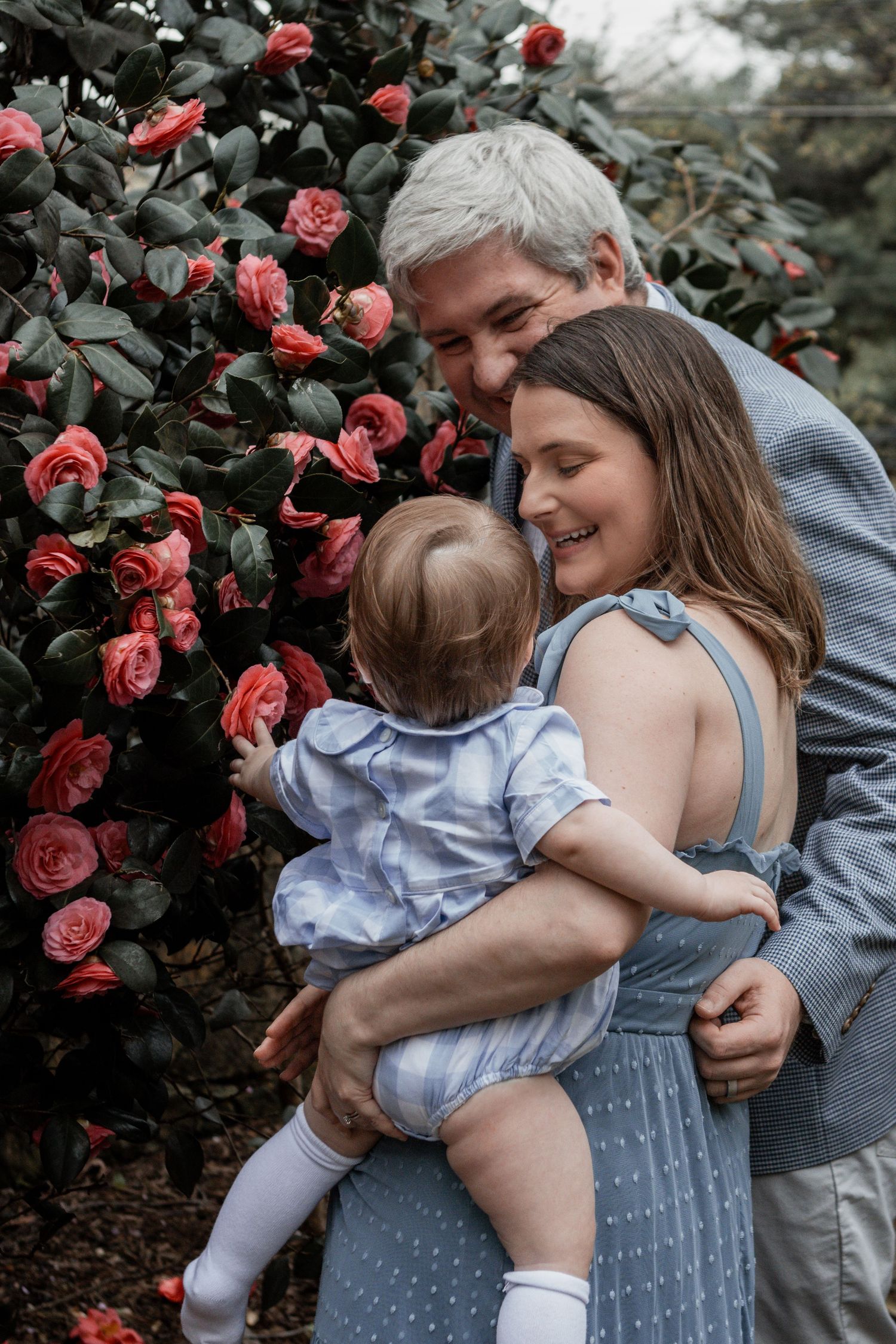 Three generations share a tender moment together by blooming pink rose bushes in a garden setting.