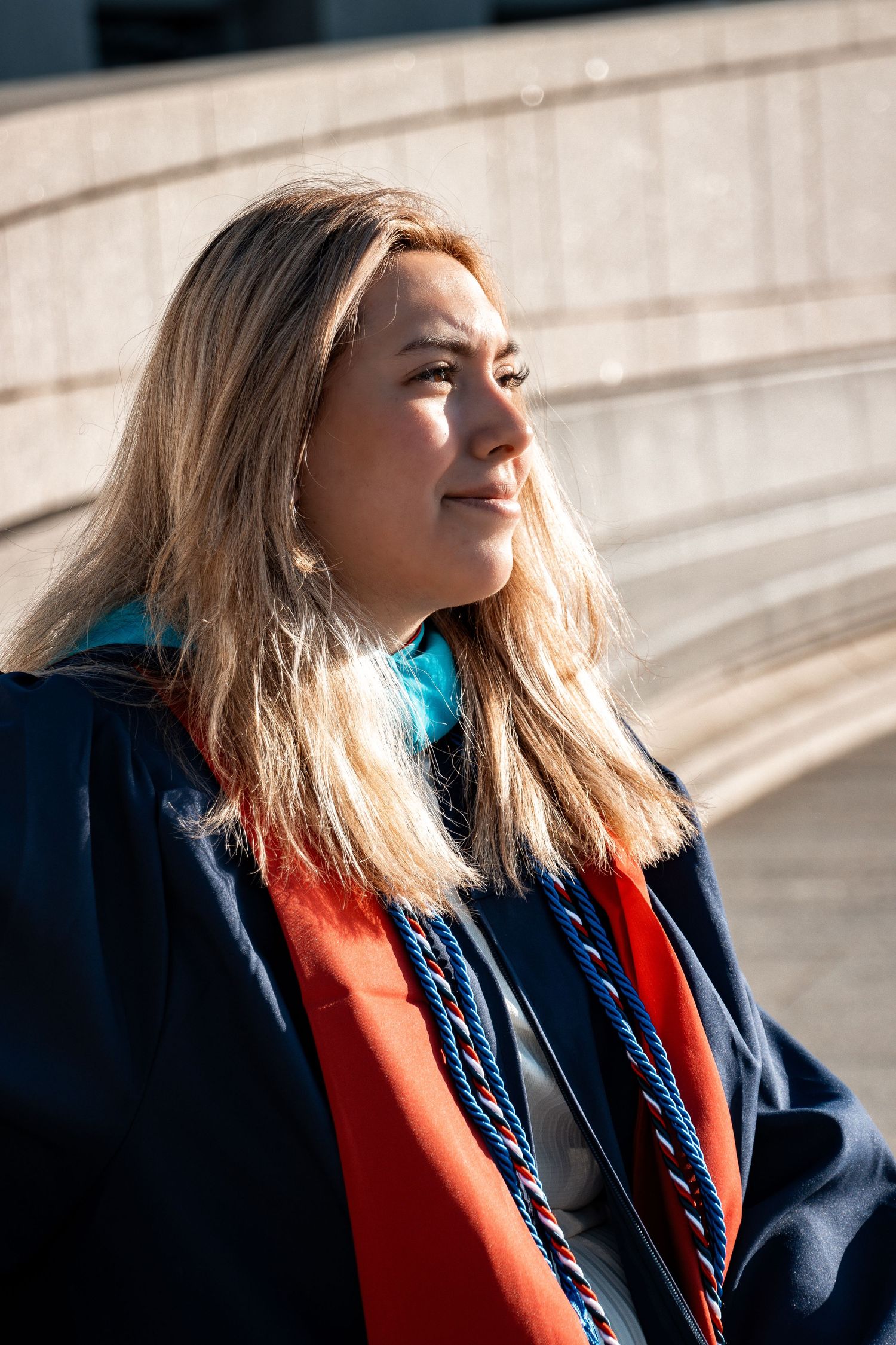 Graduate wearing blue and orange regalia poses in sunlight during graduation photoshoot.