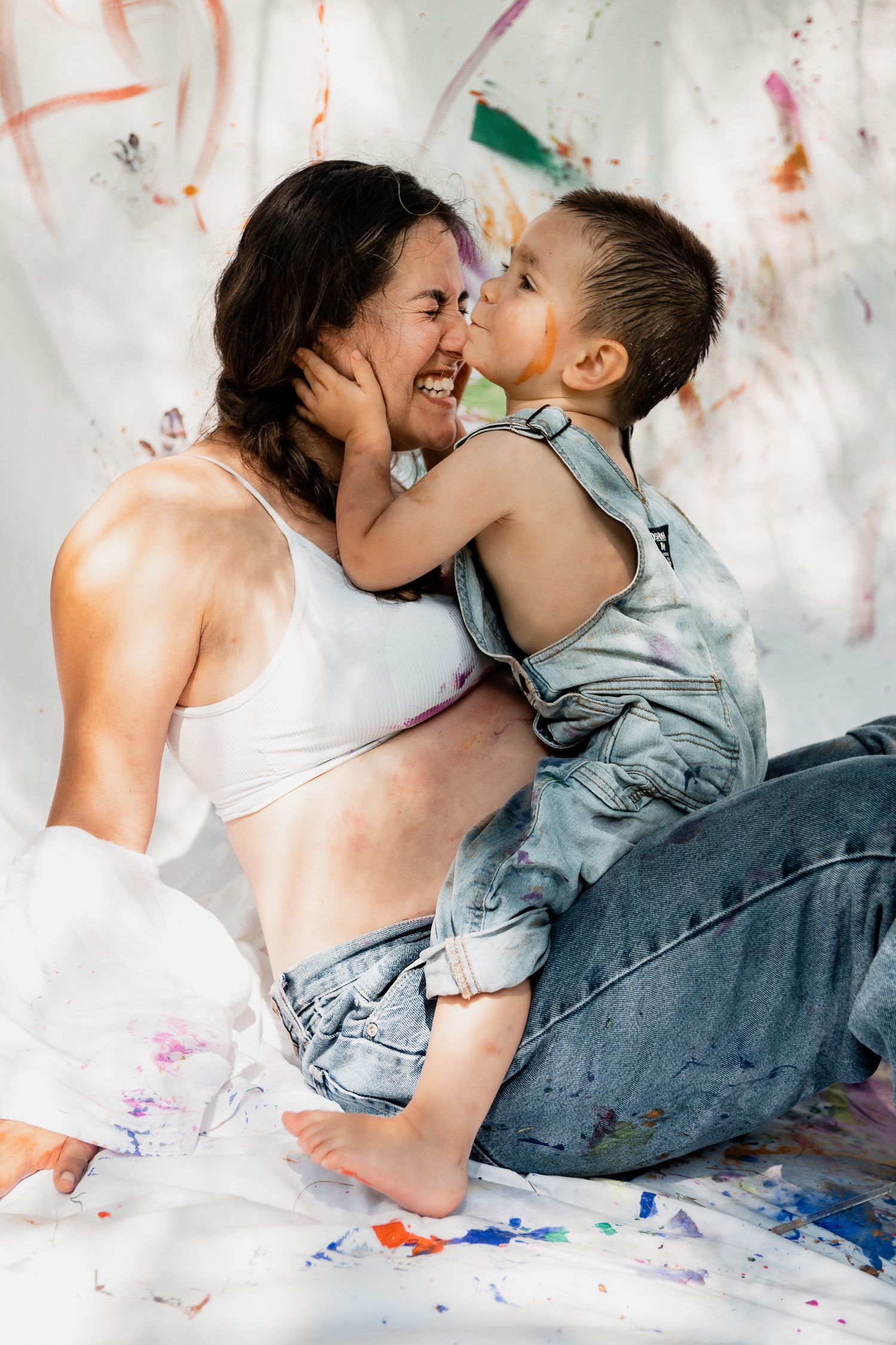 Family playing and laughing together on paint-splattered white backdrop during art session.