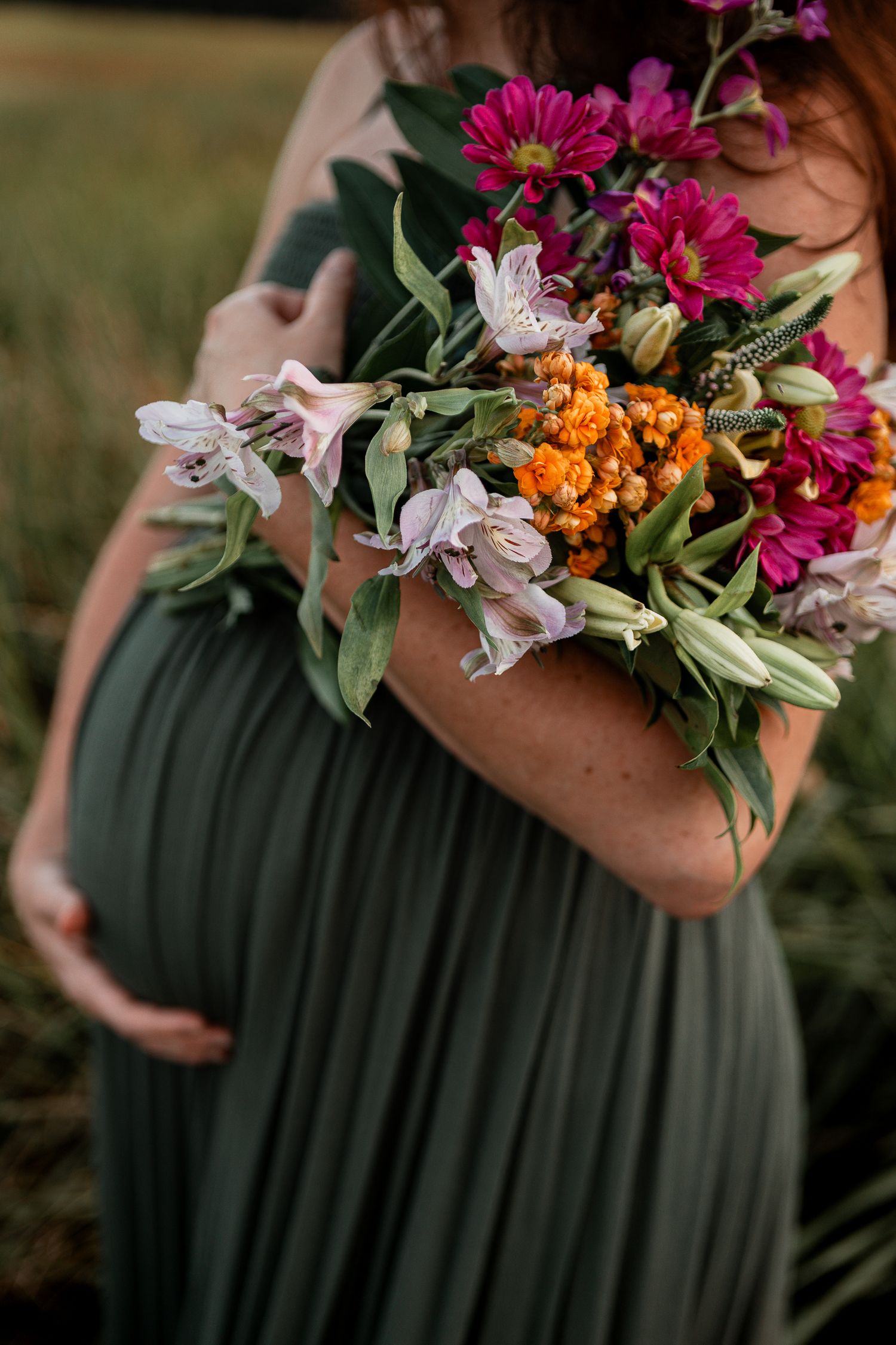 A pregnant woman in a green dress holds a vibrant bouquet with pink daisies and orange berries against a soft outdoor setting.