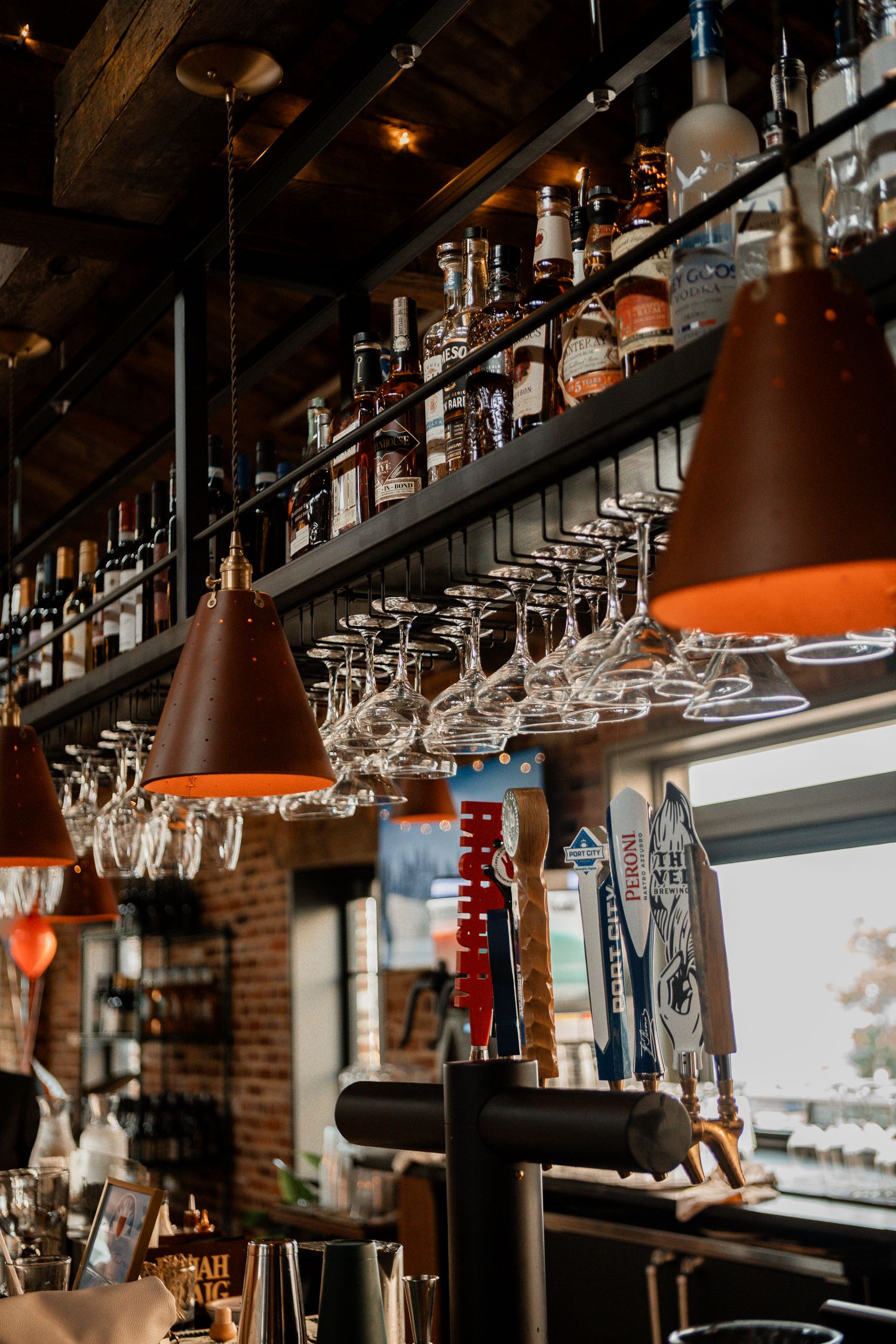 Wine glasses hanging from overhead racks in a rustic bar setting with exposed brick walls.