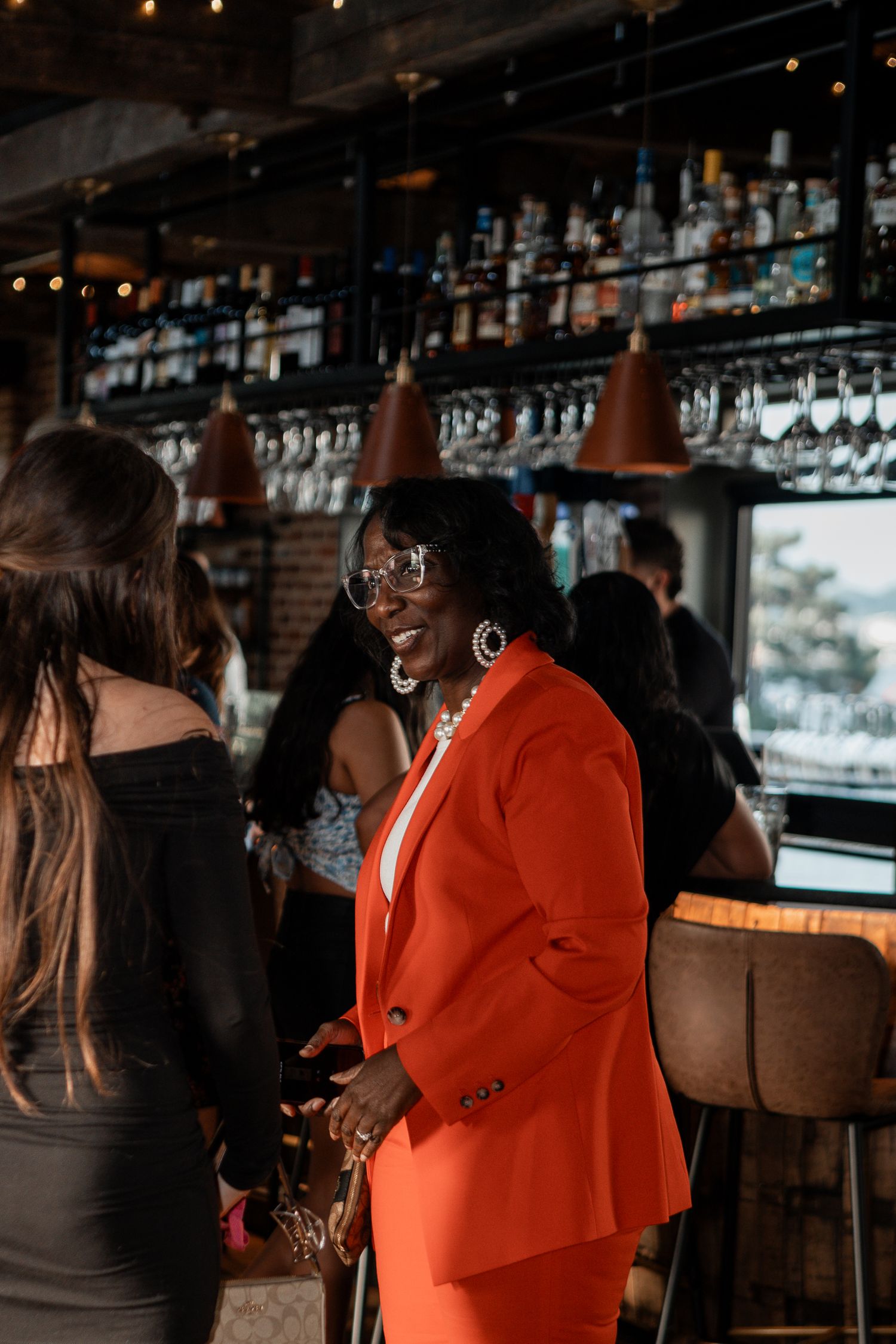 Guests socialize at a dimly lit bar with hanging pendant lights and wine glasses displayed overhead.