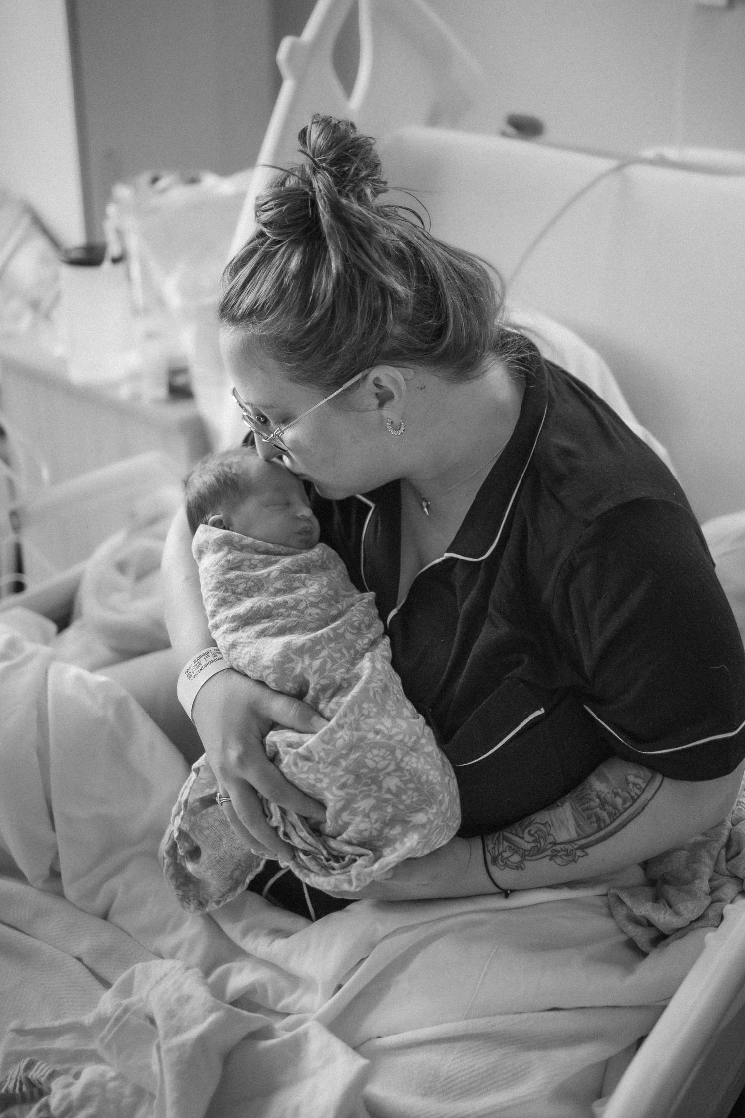 Black and white photo of a new mother in hospital gown kissing her swaddled newborn baby while sitting on a hospital bed.