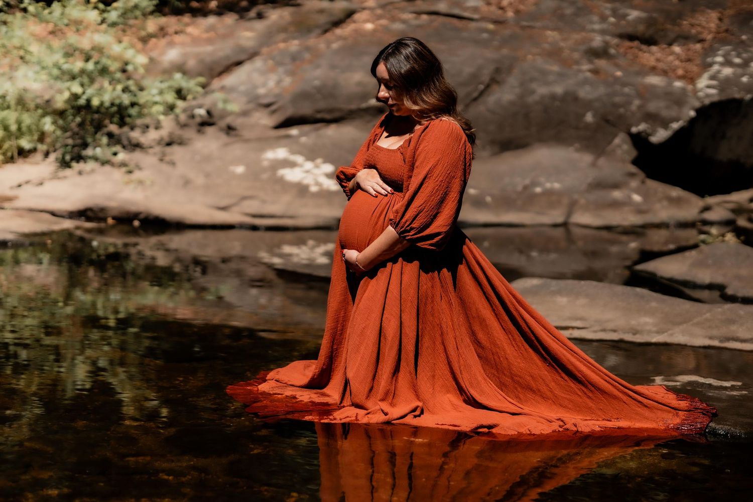 Pregnant woman in rust orange maternity gown standing in a rocky creek, cradling her baby bump.