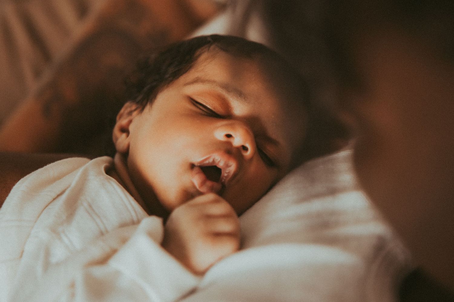 Peaceful newborn baby sleeping soundly, wearing a white onesie, with tiny hand near face in warm light.