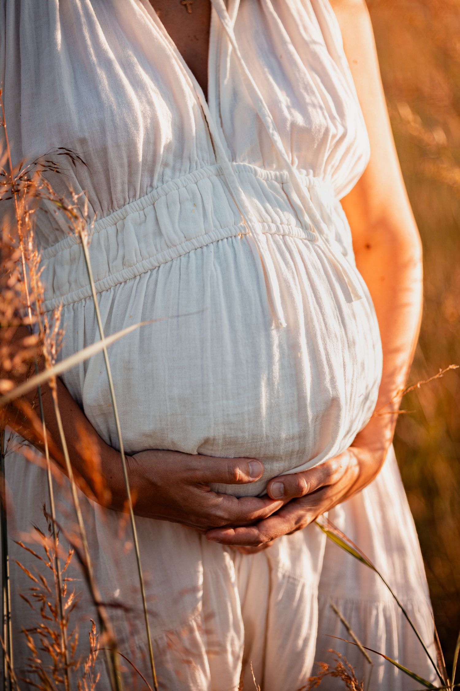 A pregnant figure in a flowing white dress cradles their belly while standing among tall grass at golden hour.